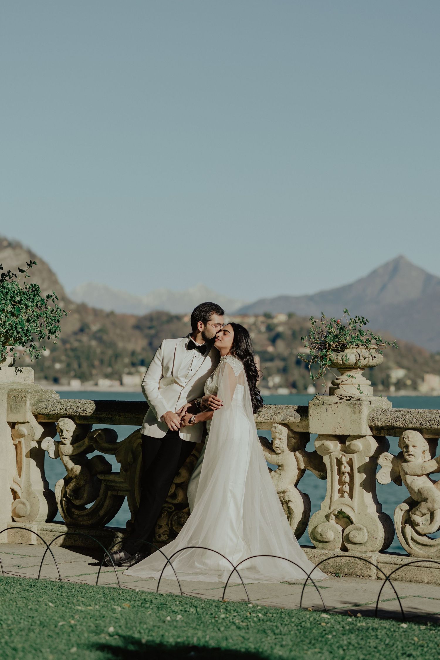 Bride and groom share a romantic kiss by ornate stone balustrade with lake and mountains backdrop.