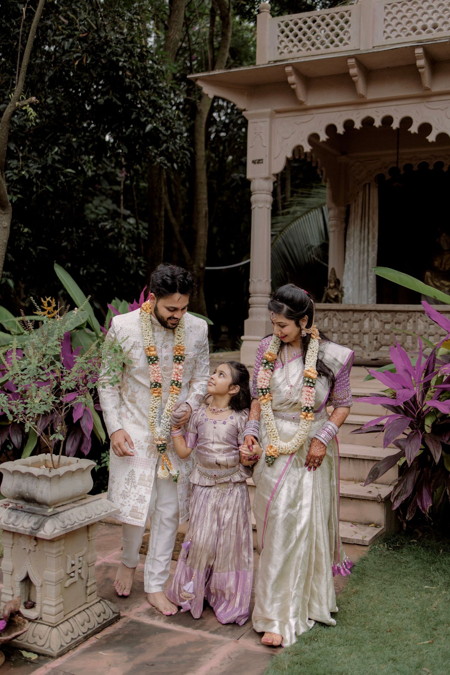Indian couple in traditional wedding attire with flower garlands walking with young girl in garden.