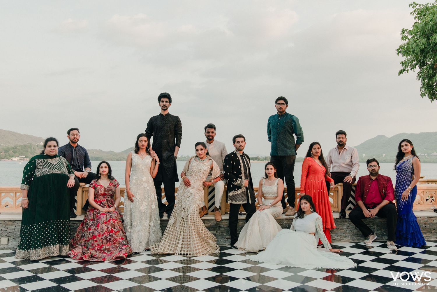 Large Indian wedding group photo outdoors by a lake, guests in colorful traditional attire on a checkered floor.