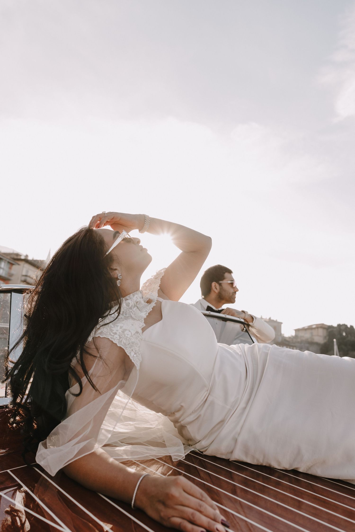 Bride in white lace dress reclines on wooden boat deck while groom sits beside her during golden hour.