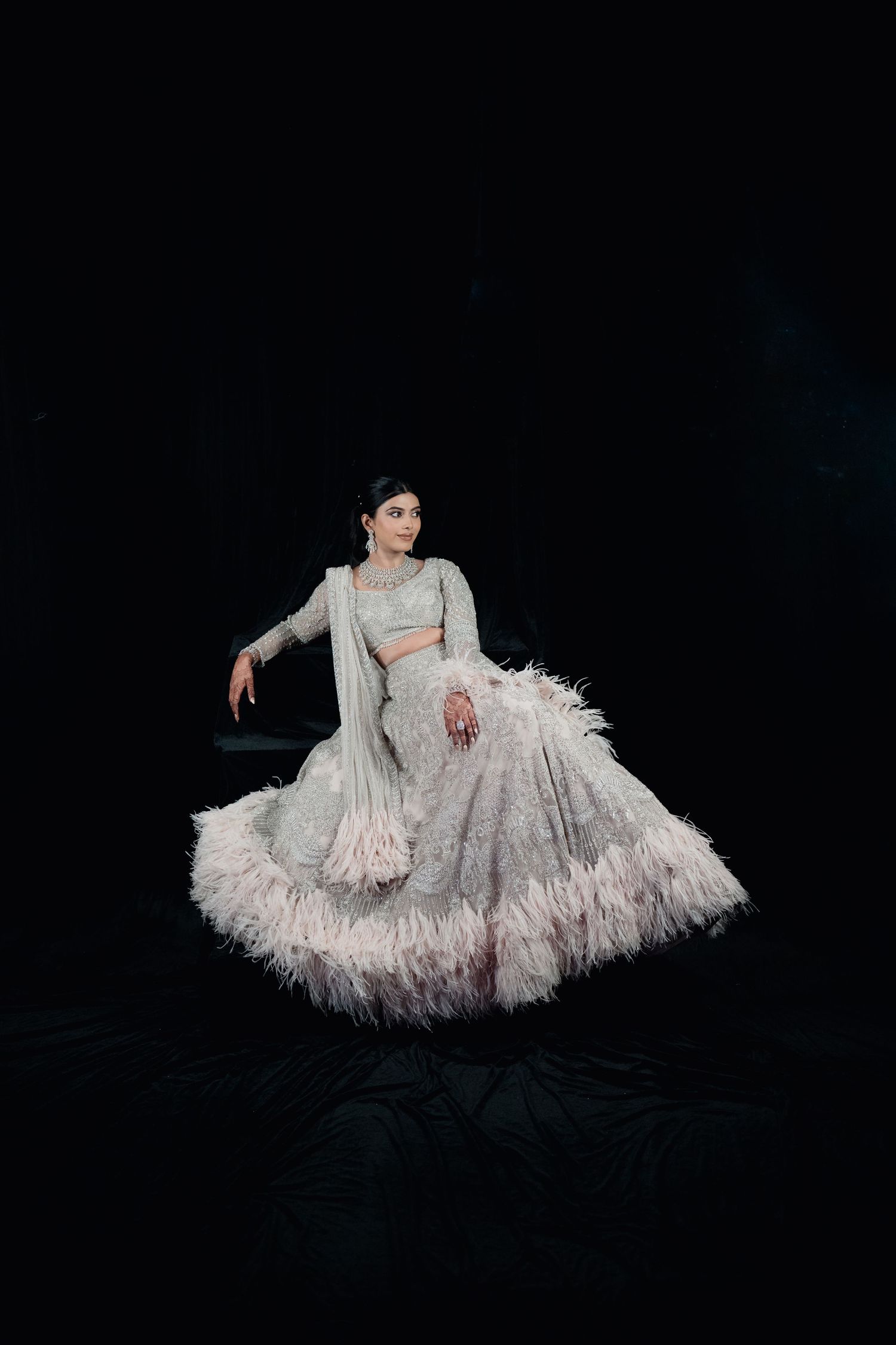 Woman in silver embroidered lehenga with feather hem trim poses gracefully against dark studio background.