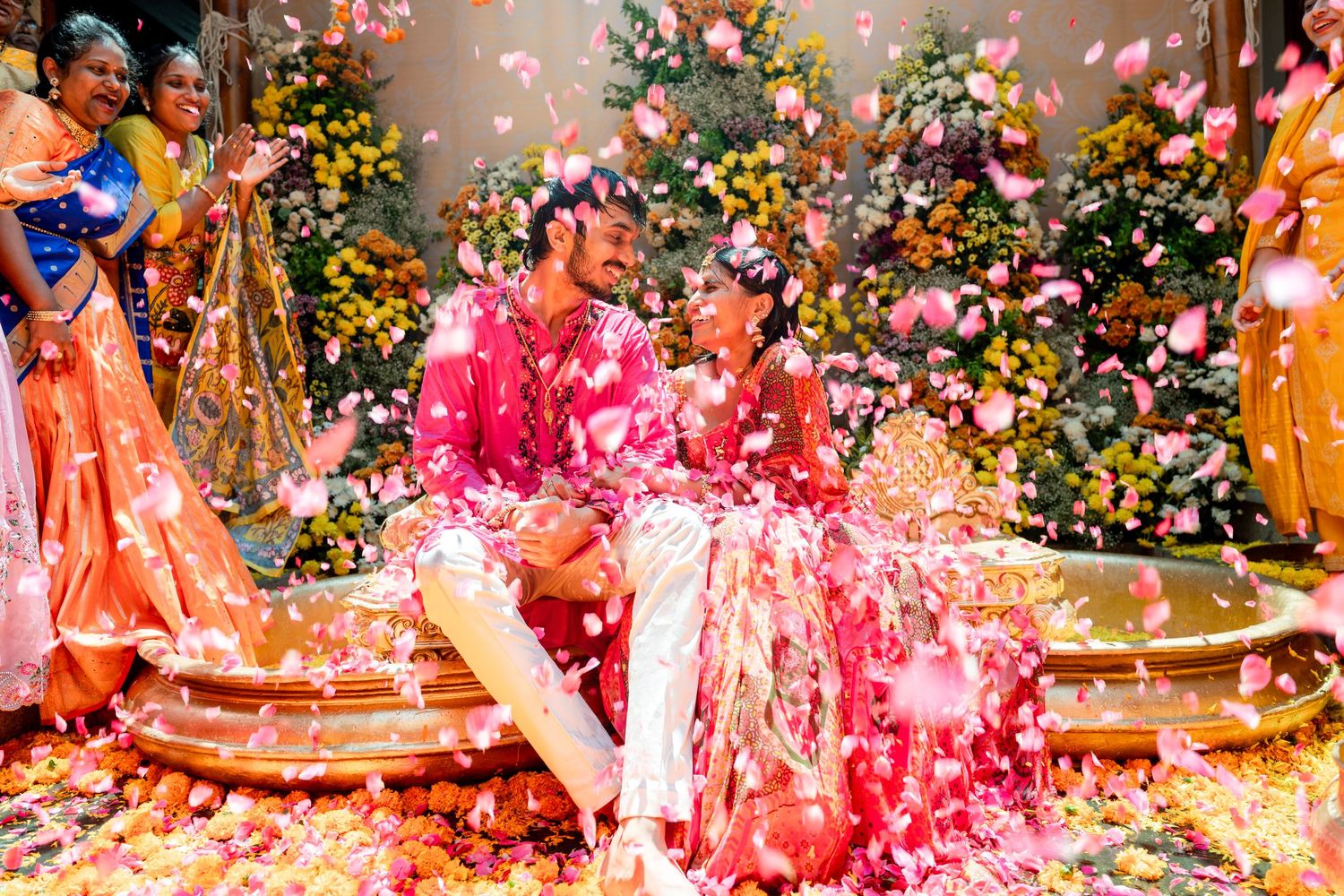 Joyful Indian couple showered with pink flower petals during vibrant wedding haldi ceremony.