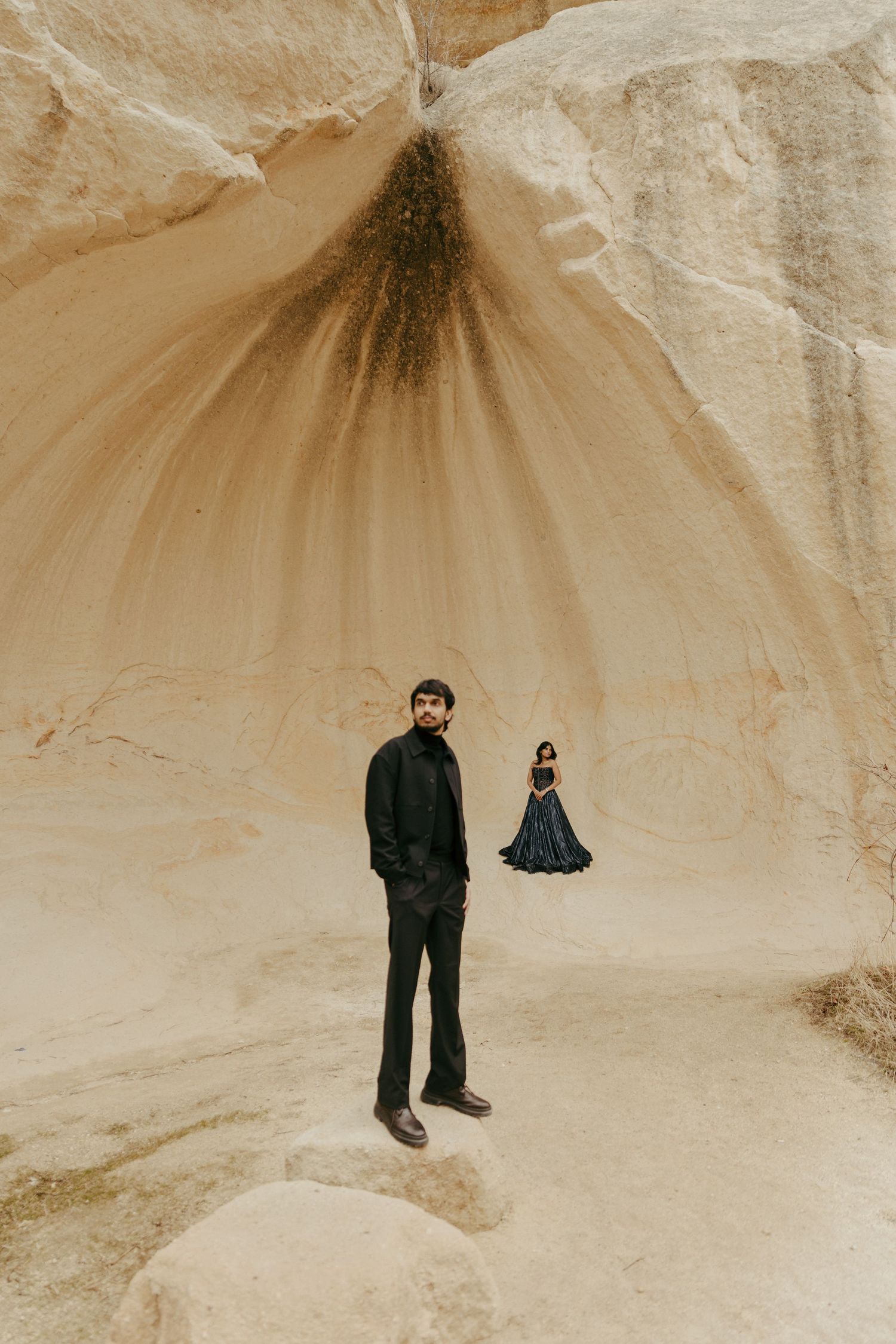 A man in black stands in a sandy canyon while a woman in a teal gown poses in the background.