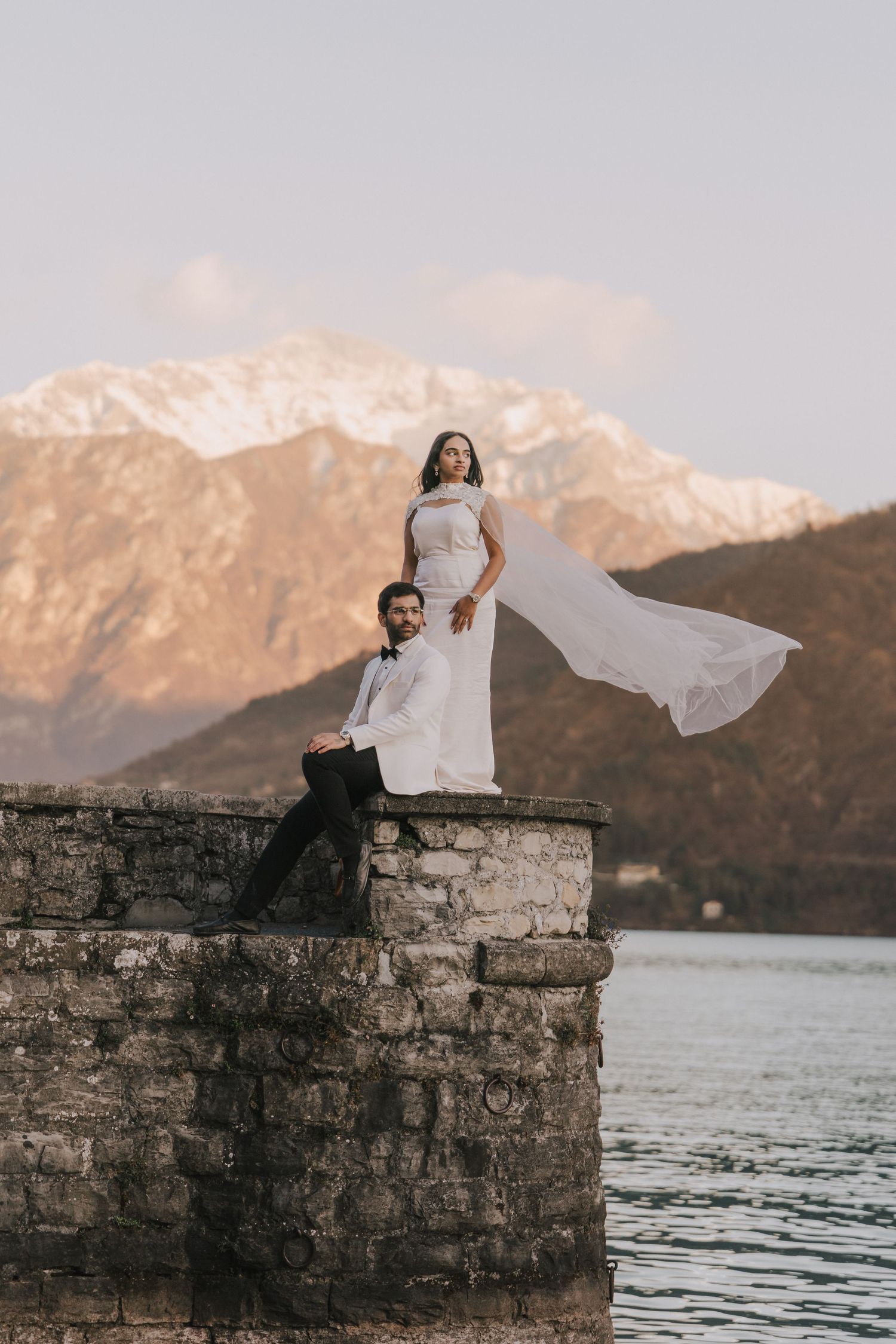 Bride and groom posing on stone wall by lake with snow-capped mountains at sunset, wedding dress flowing in wind.