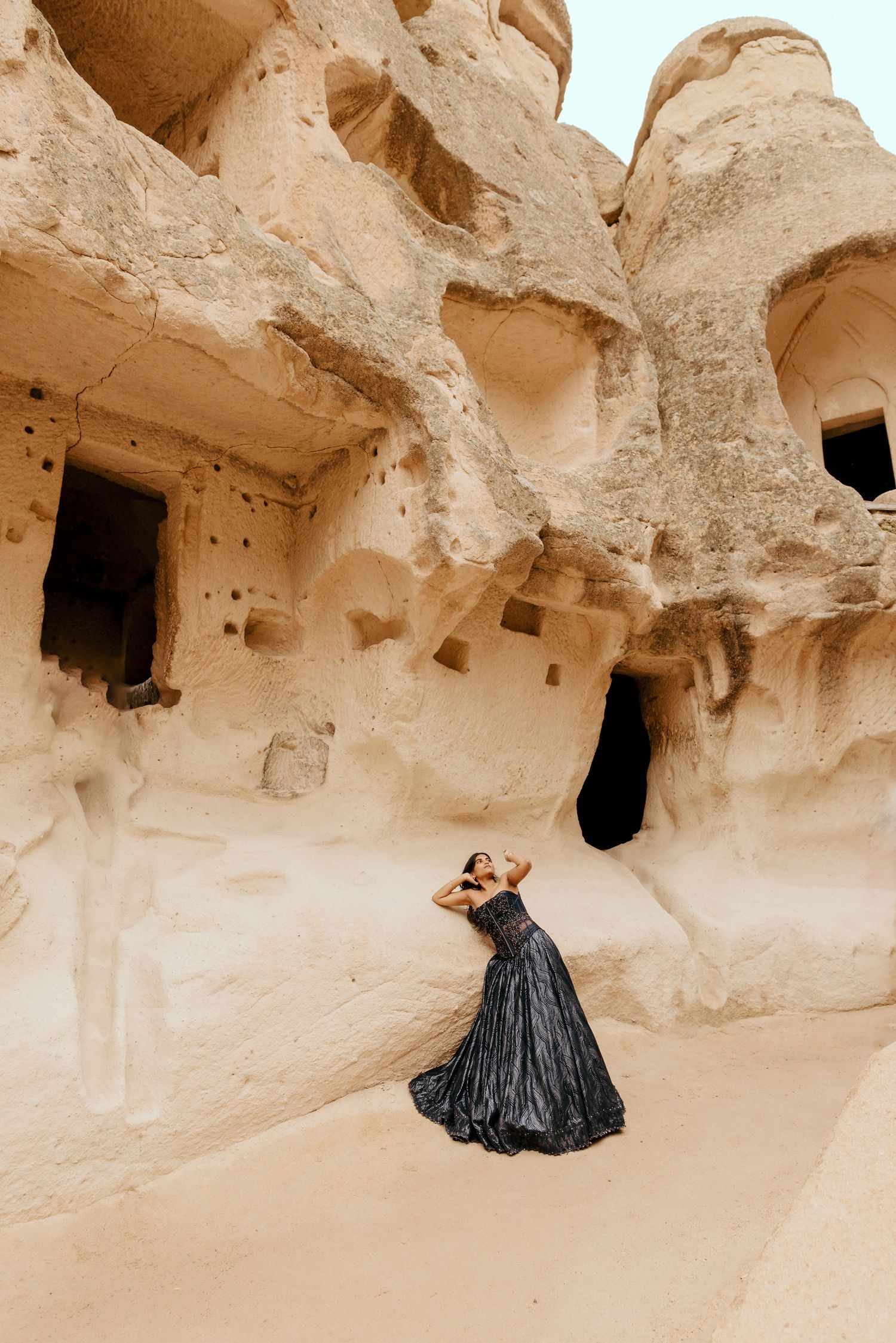 Woman in black gown poses before ancient cave dwellings carved into sandstone cliffs in Cappadocia, Turkey.