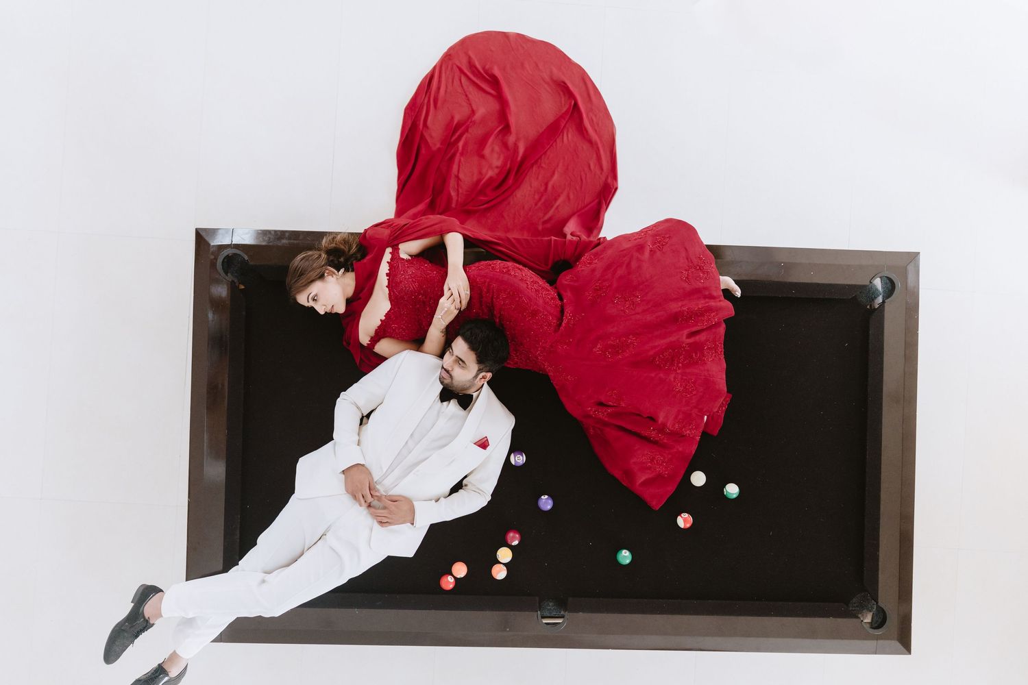Aerial view of a couple in white suit and red gown lying on a black pool table with colorful balls.