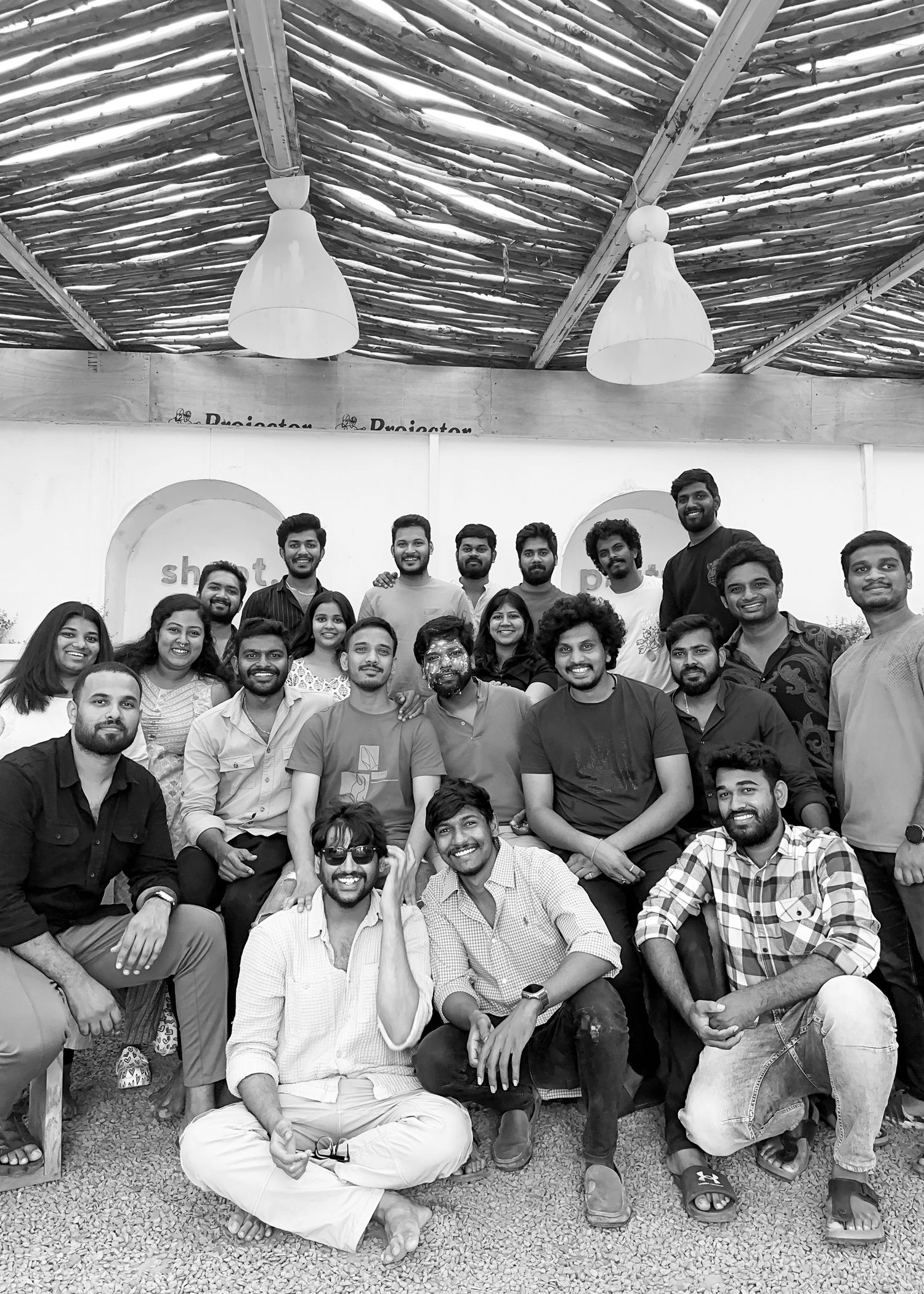 Black and white group photo of young adults posing together indoors under a thatched roof ceiling.