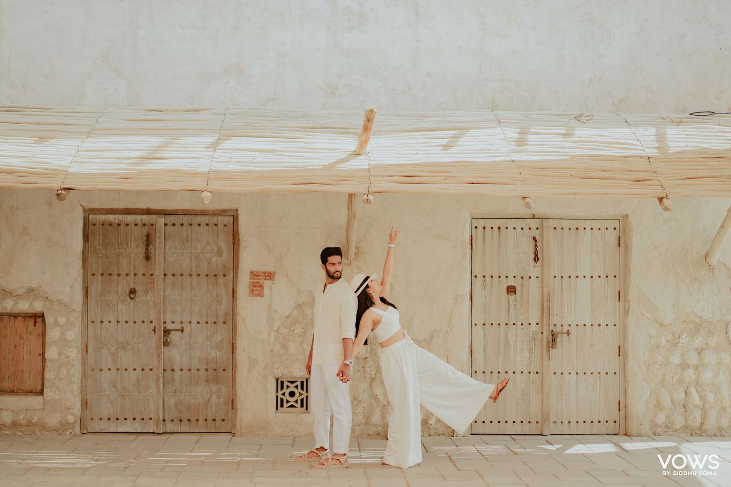 Couple in white outfits posing near rustic wooden doors in a desert setting for a pre-wedding shoot.