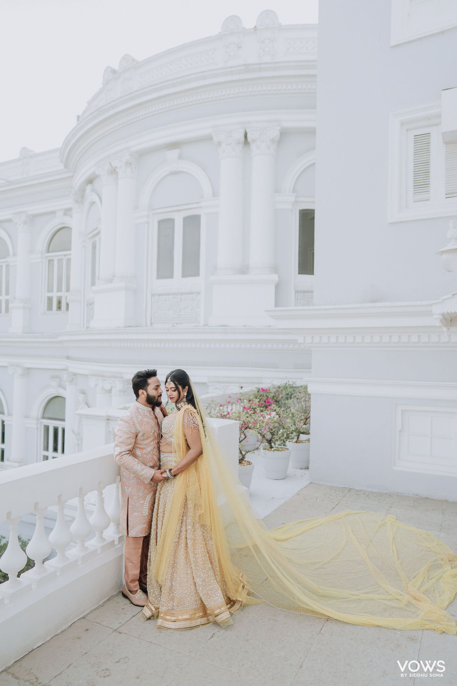 Indian bride in yellow lehenga with flowing dupatta poses with groom in pink sherwani on white palace balcony.