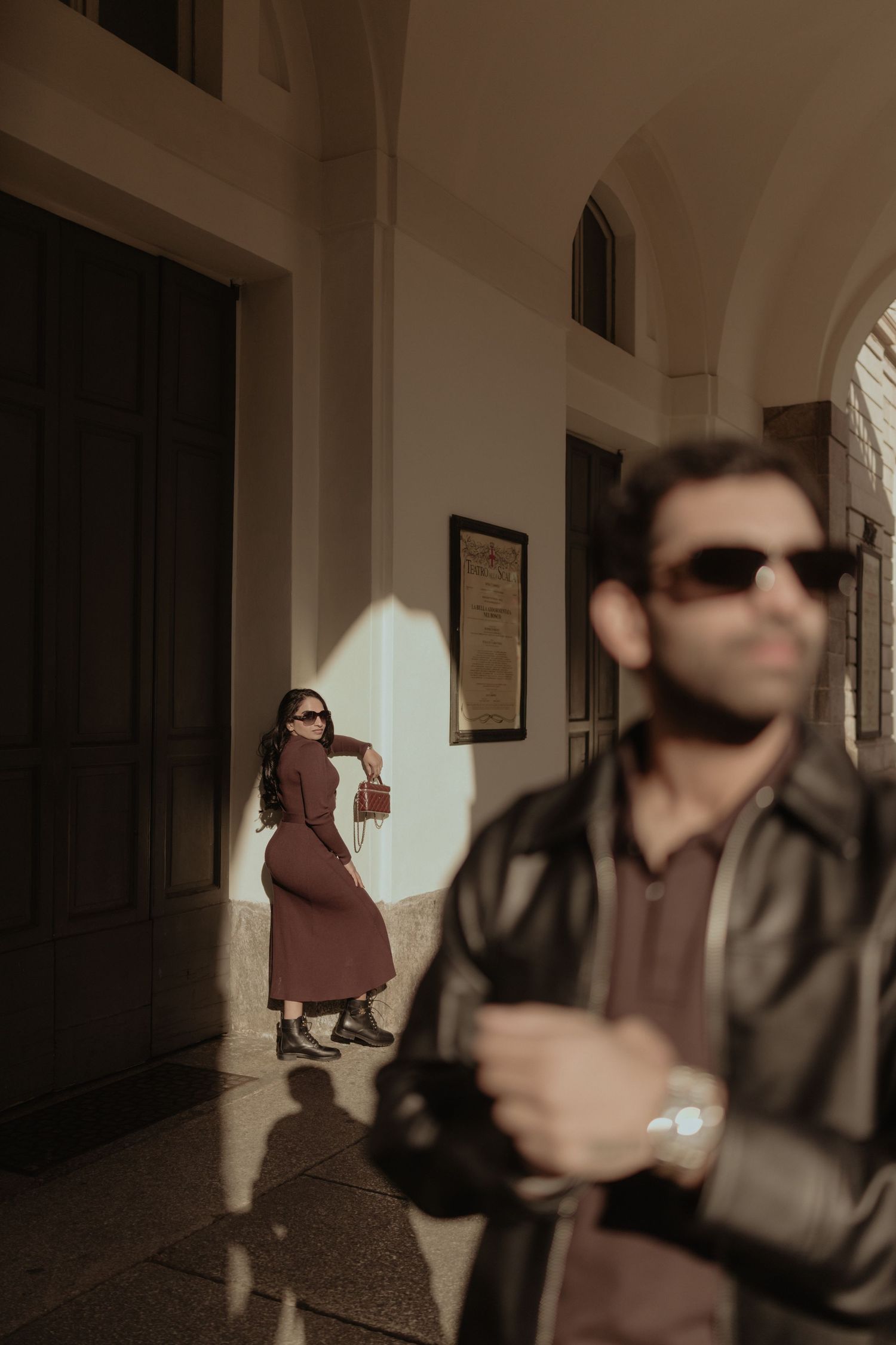 Stylish couple in sunglasses pose in sunlit archway corridor, woman in burgundy dress with handbag.