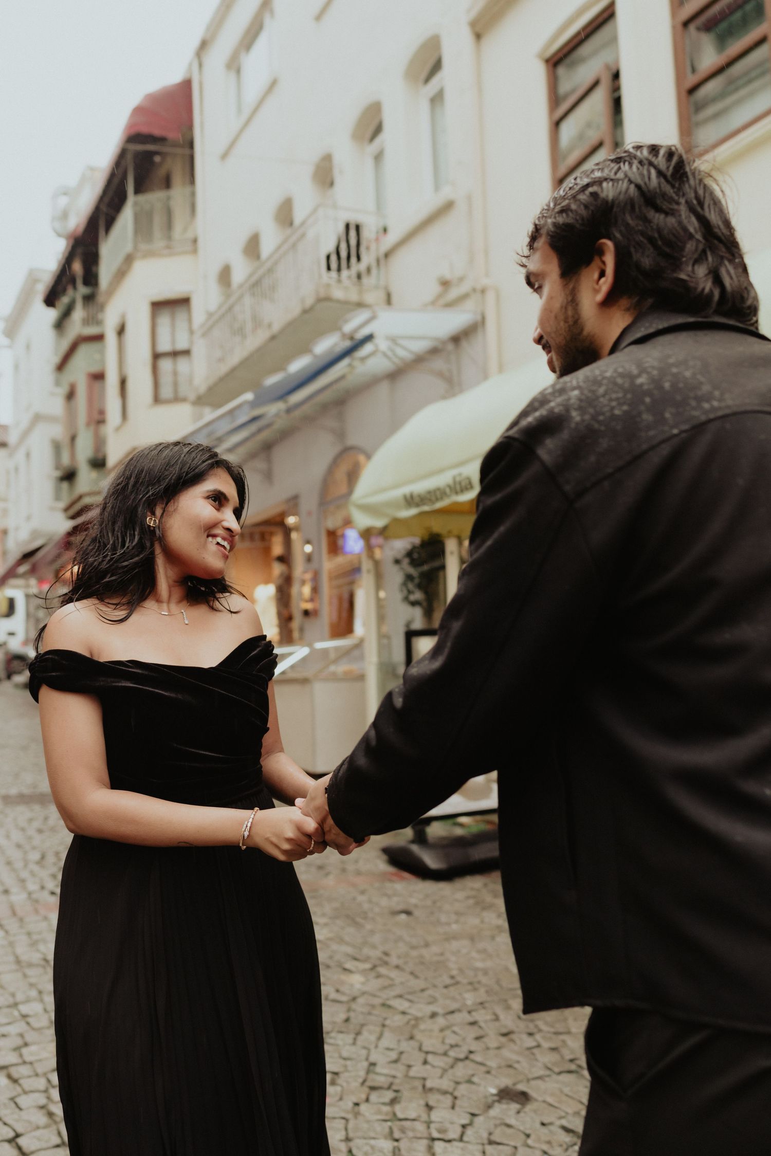 Couple in black outfits holding hands and smiling on a cobblestone street in a European town.