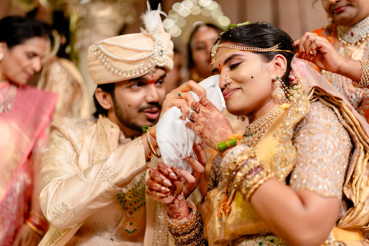 A groom tenderly feeds his bride during a traditional South Indian wedding ceremony in golden bridal attire.