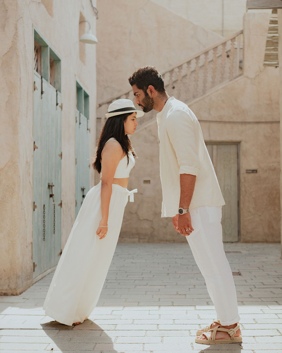 Couple in matching white outfits facing each other in a sunlit stone alleyway, sharing a romantic moment.
