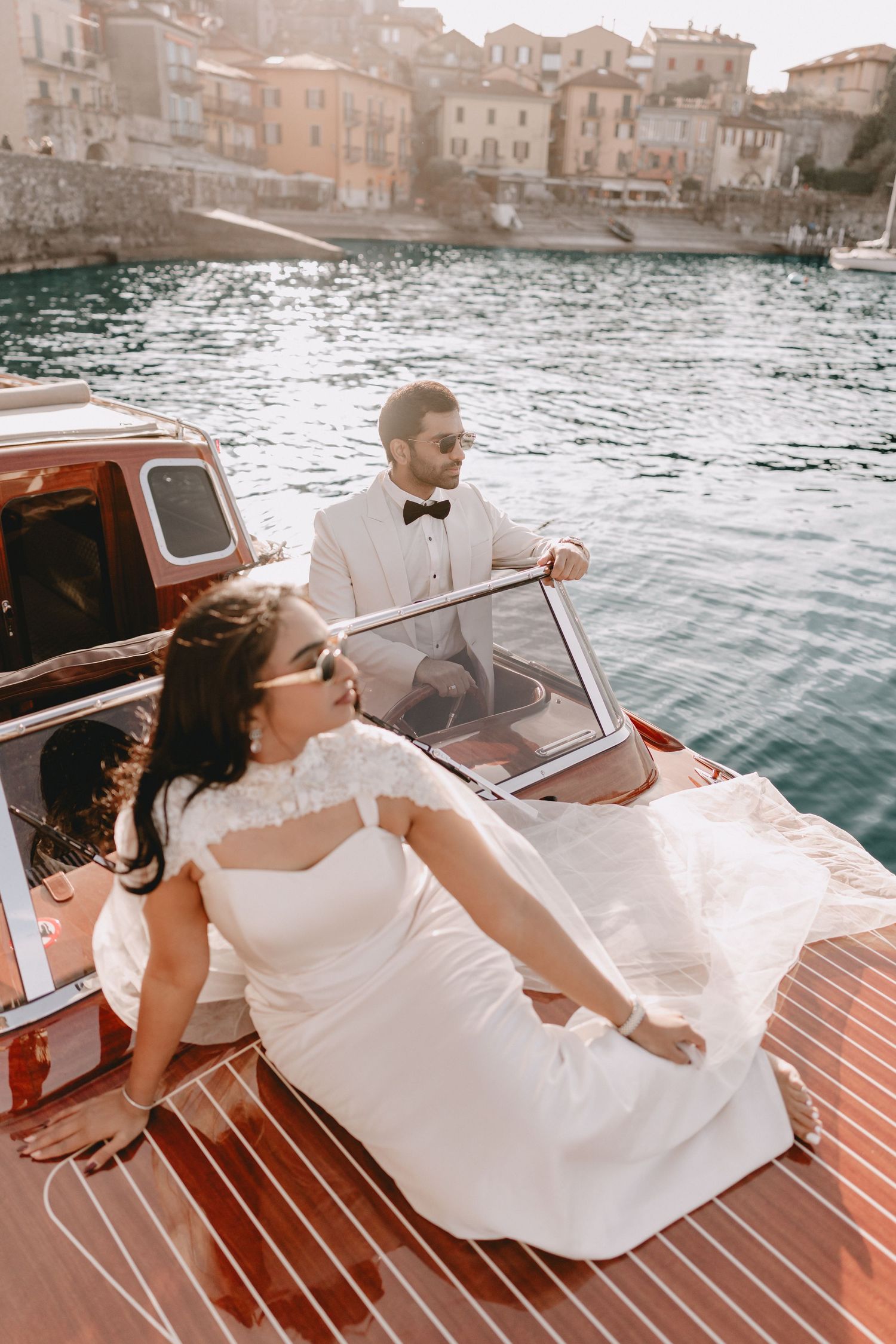 Bride in white gown and groom in tuxedo on a wooden boat with Italian village backdrop on a sunny day.