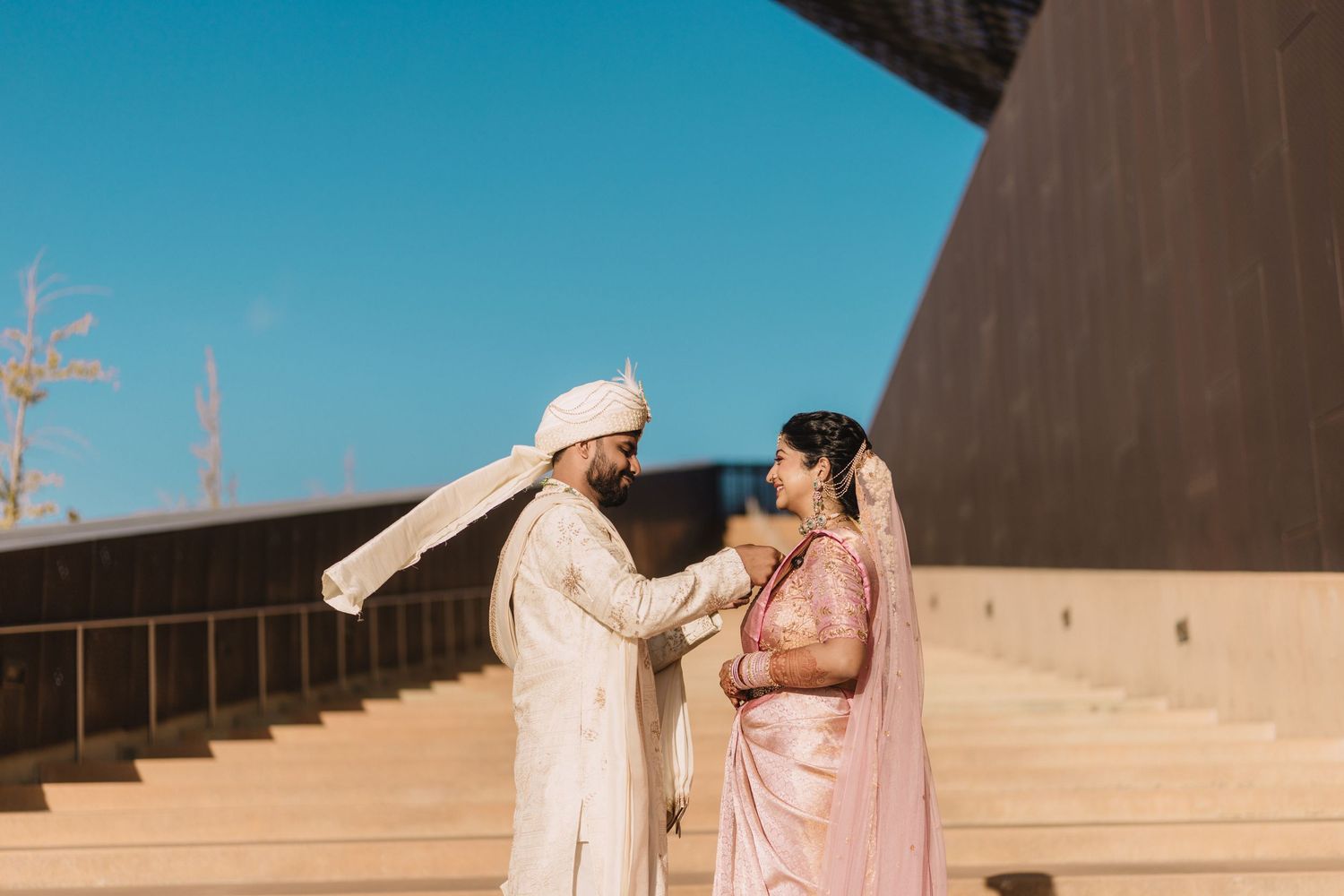 Indian bride in a pink saree and groom in a white sherwani and turban share a tender moment on modern stone steps under a clear blue sky.
