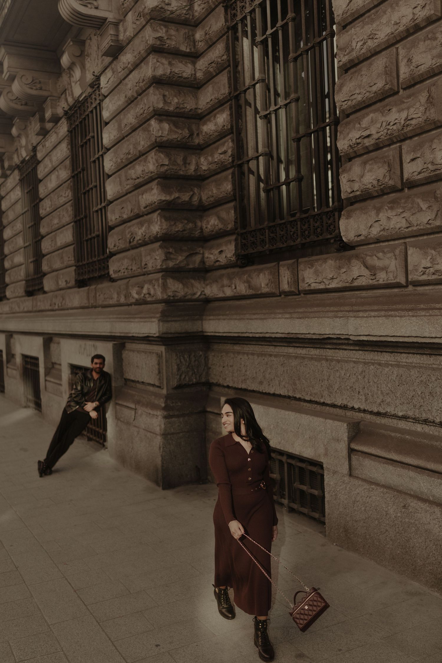 Woman in burgundy dress with handbag walks past historic stone building as man leans against wall behind her.