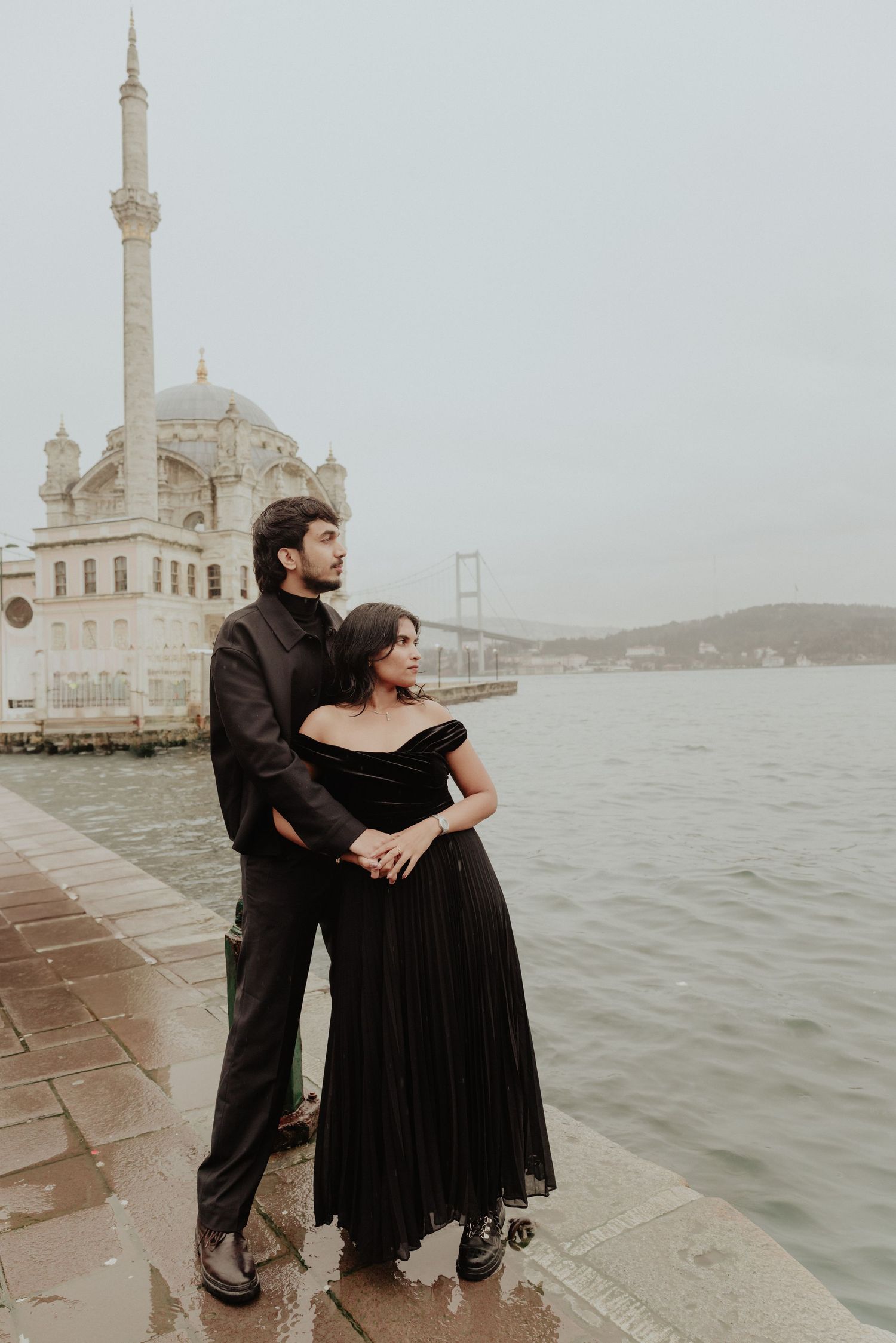 Romantic couple in matching black outfits embracing by the Bosphorus with Ortakoy Mosque in the background, Istanbul.