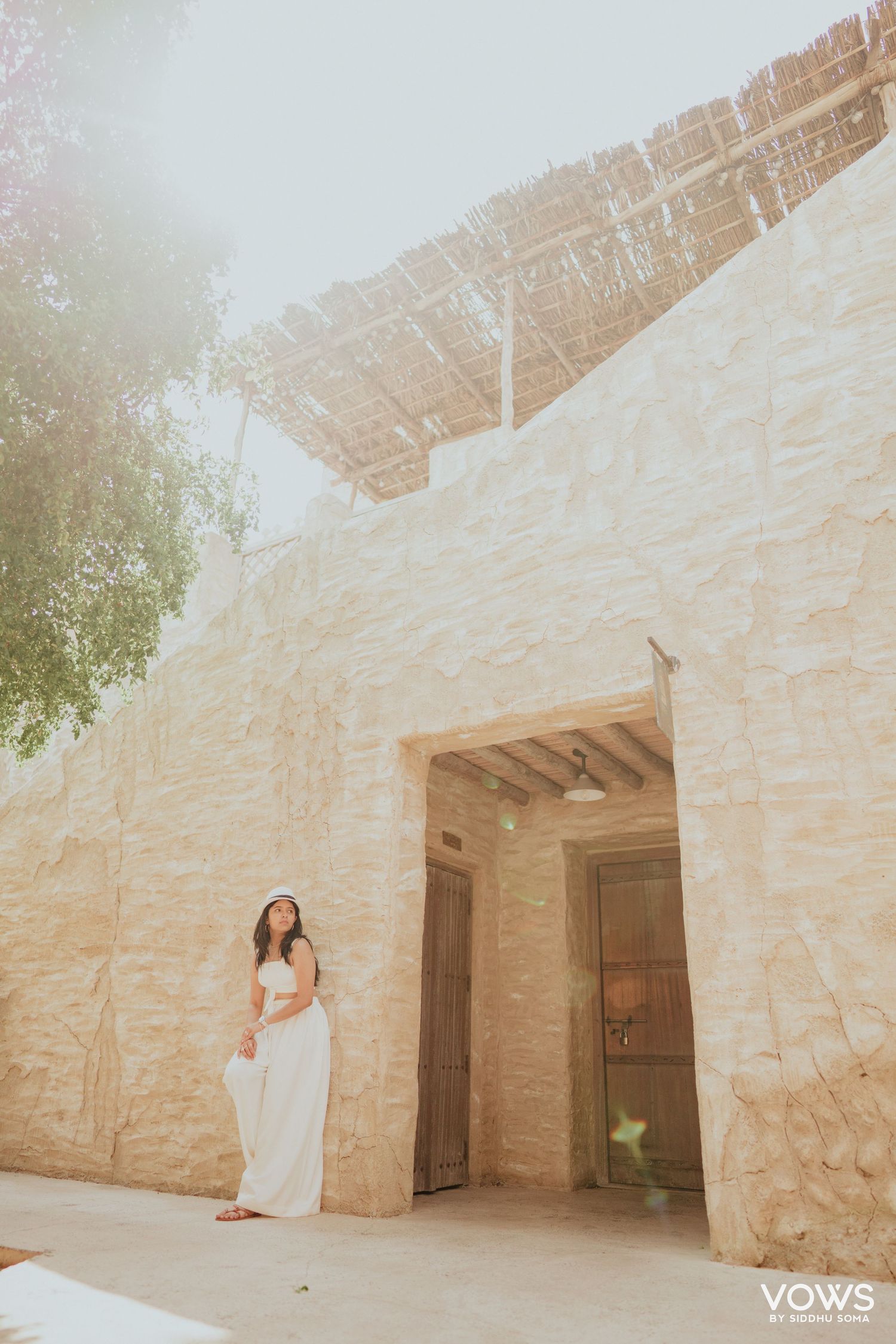 Woman in white dress posing by rustic stone wall entrance with thatched roof at a desert resort.