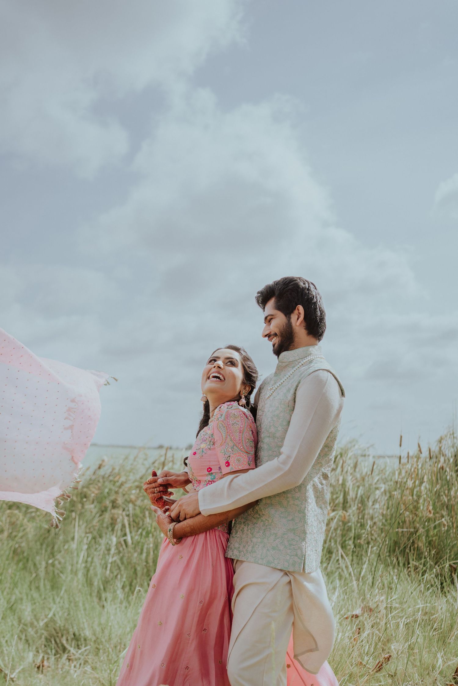 Smiling couple in traditional Indian attire embracing in a grassy field under a cloudy sky, with a pink dupatta fluttering in the breeze.