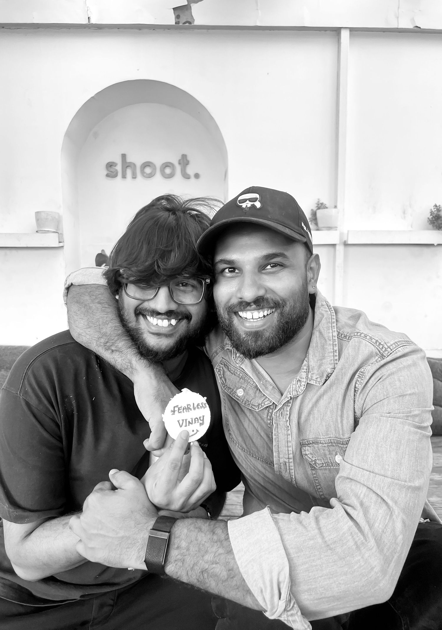 Two bearded men smiling and posing together indoors, one holding a badge, in black and white photo.