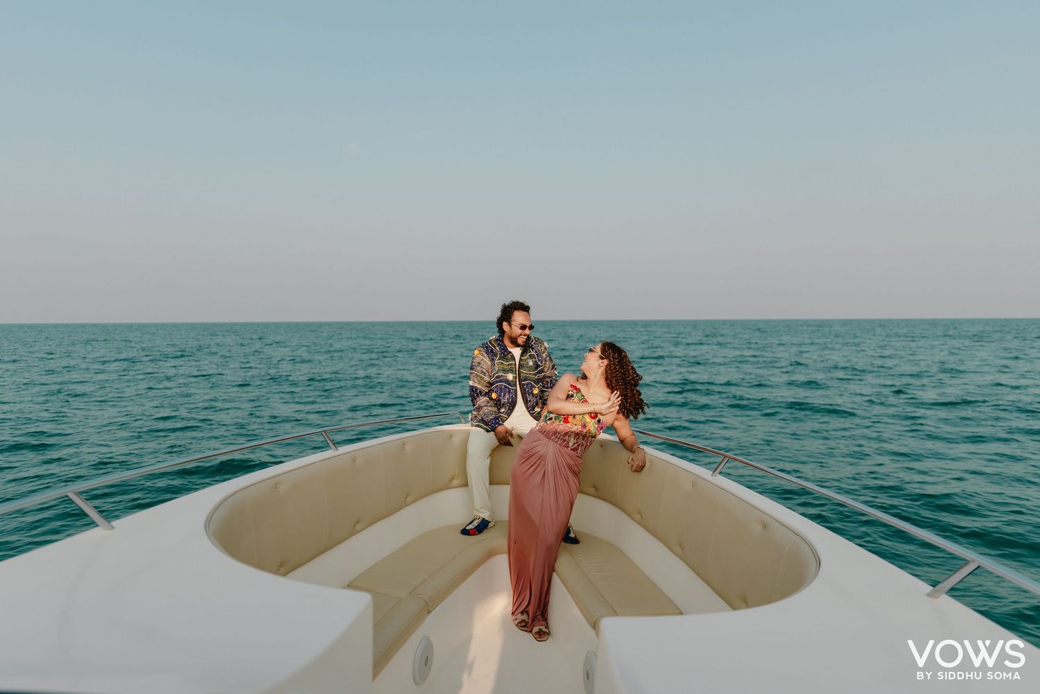 Couple celebrating on the bow of a luxury boat on calm turquoise ocean waters at sunset.