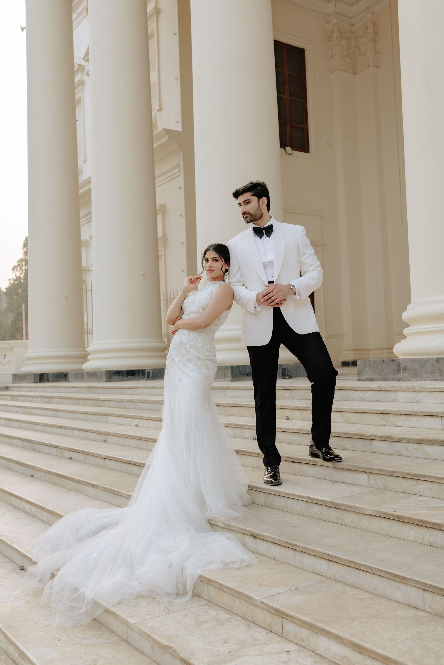 Bride in white mermaid gown and groom in white tuxedo pose on marble steps of elegant columned venue.