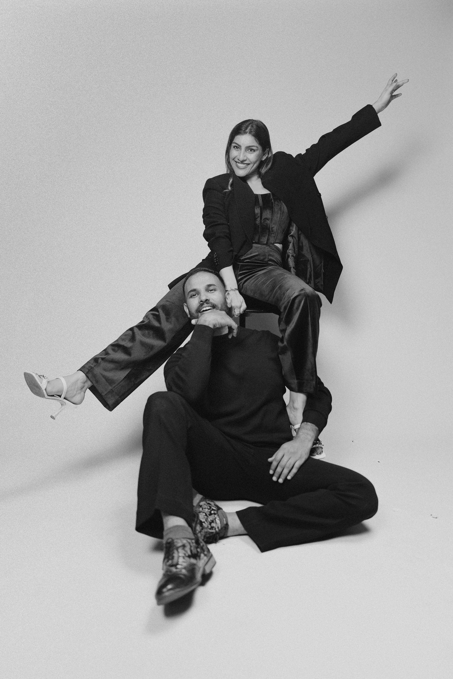 Black and white photo of two stylish people posing playfully, one sitting on the other's shoulders, both wearing elegant dark outfits.