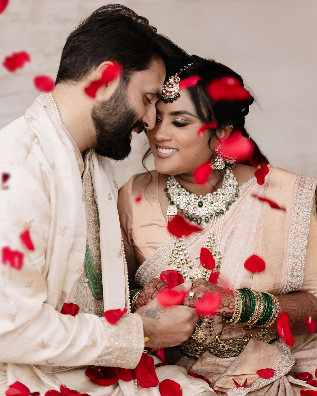 Indian bride and groom in traditional attire share a romantic moment surrounded by falling red rose petals.