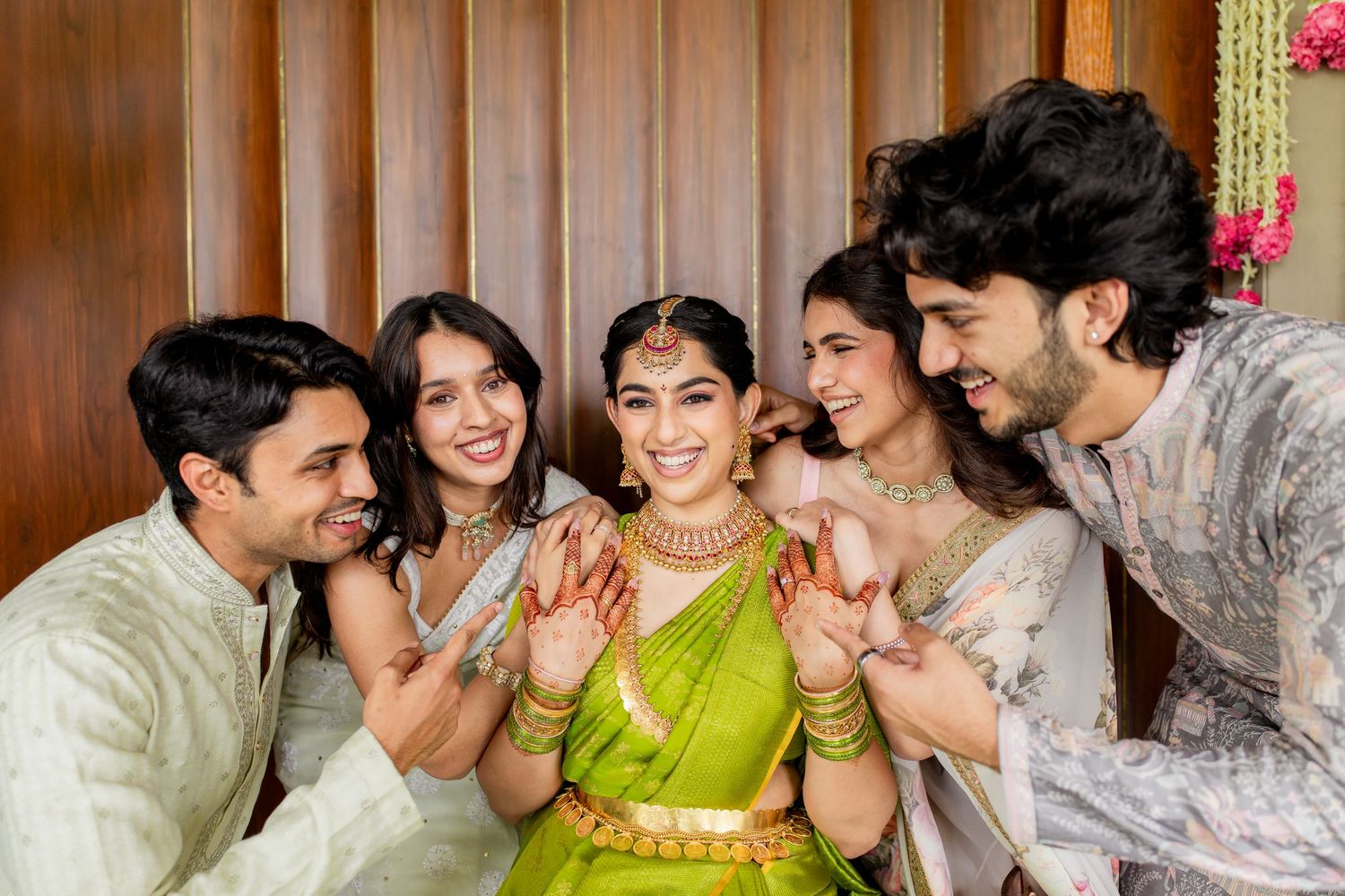 Bride in green saree with mehndi hands surrounded by smiling friends in traditional Indian attire.