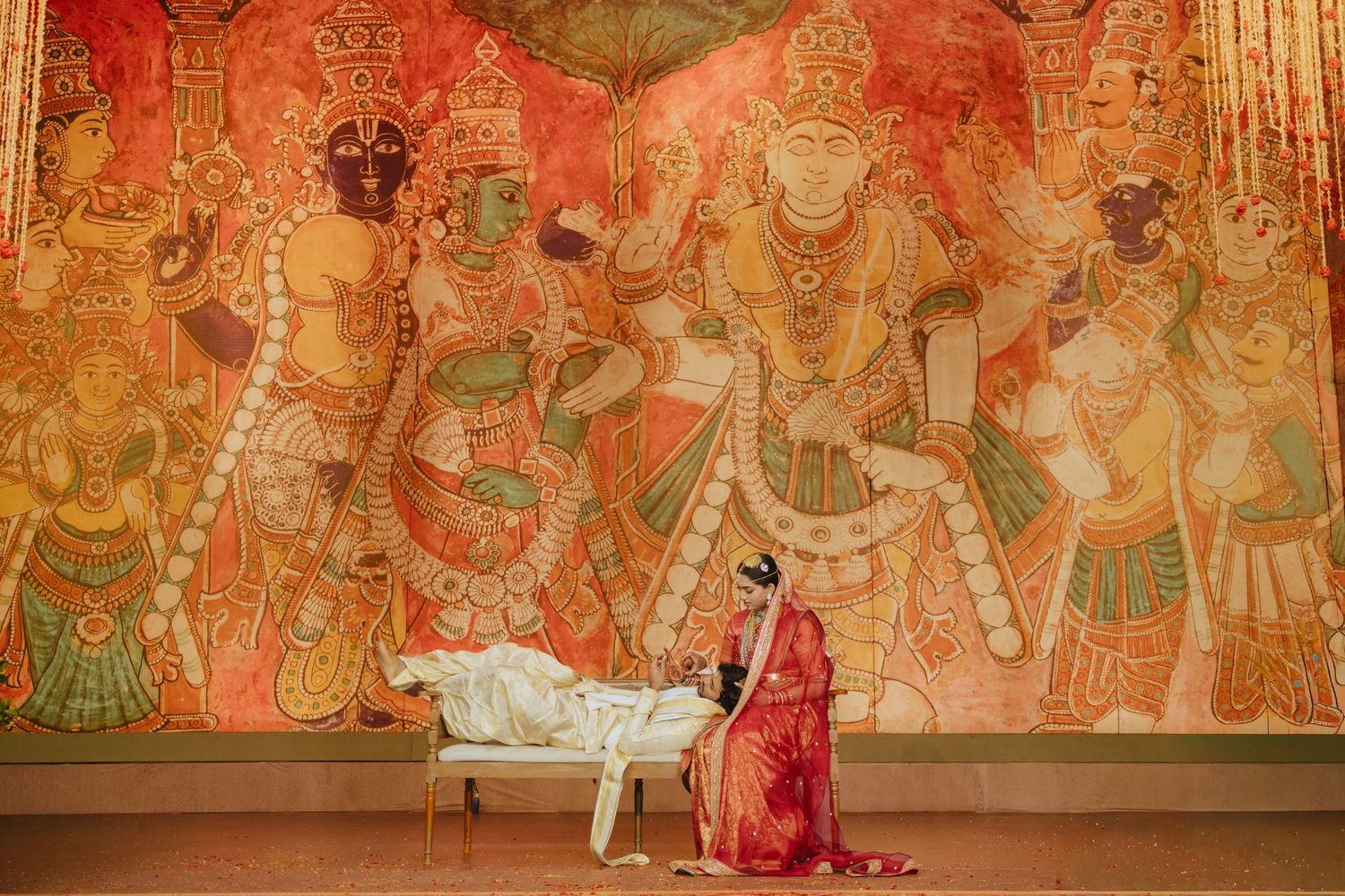 Woman in red saree performing traditional Indian ritual before colorful Hindu temple mural painting backdrop.