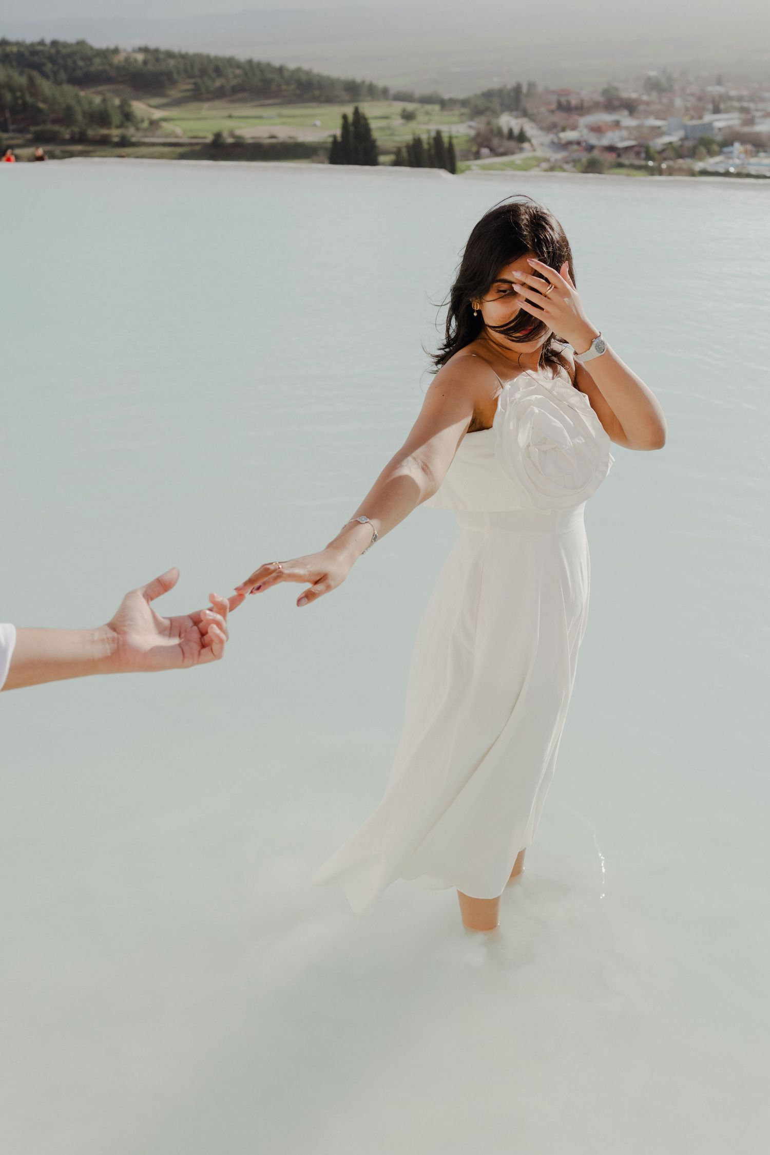 Bride in white dress wading in turquoise water holding outstretched hand with scenic landscape behind.