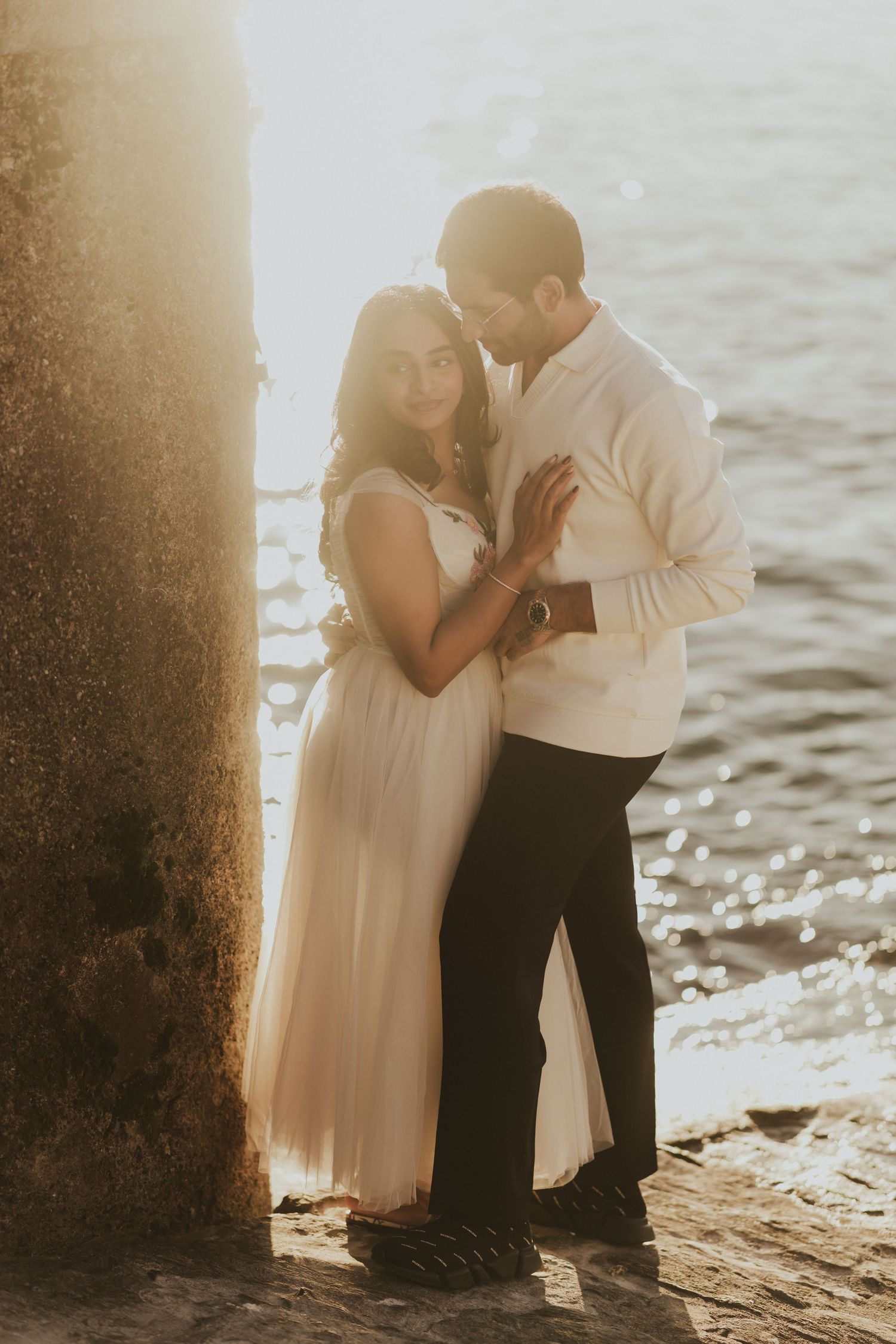 Couple embracing by sunlit waterfront, woman in white dress and man in white shirt holding hands at golden hour.