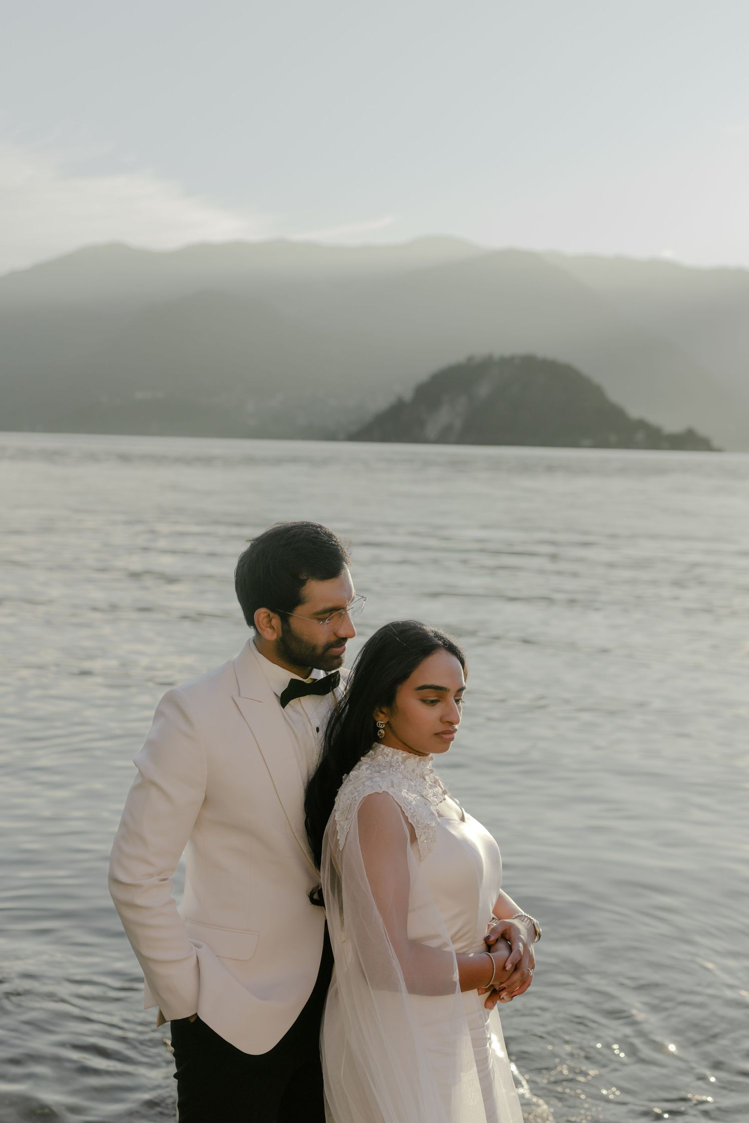 Bride and groom in wedding attire pose by a serene lake with misty mountains in the background.