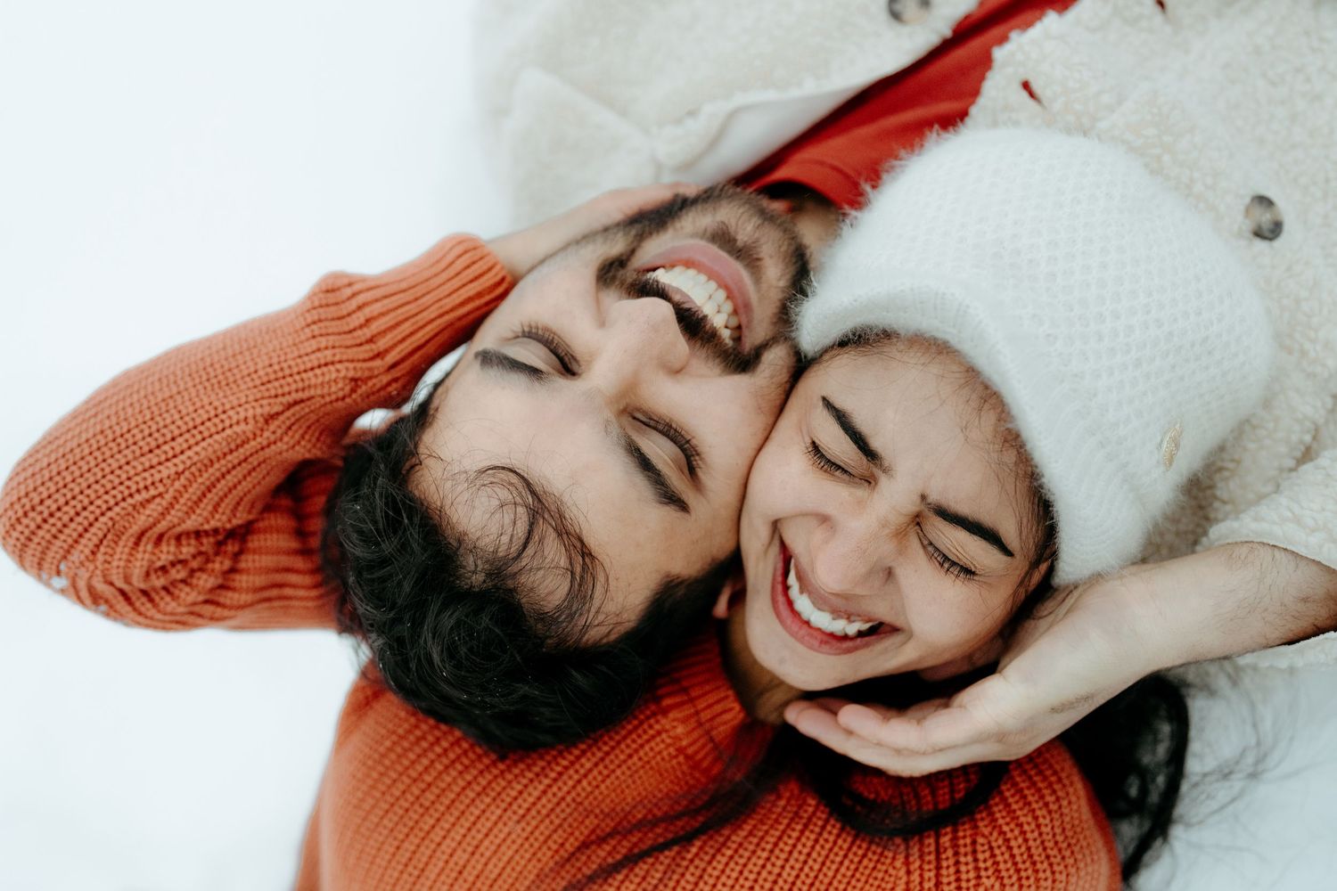 Happy couple laughing together in winter snow, wearing cozy orange and white sweaters, sharing a joyful moment outdoors.