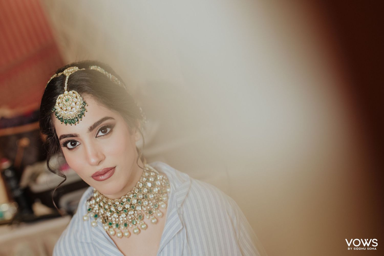 Indian bride in kundan necklace and maang tikka poses elegantly in a striped shirt during bridal photoshoot.