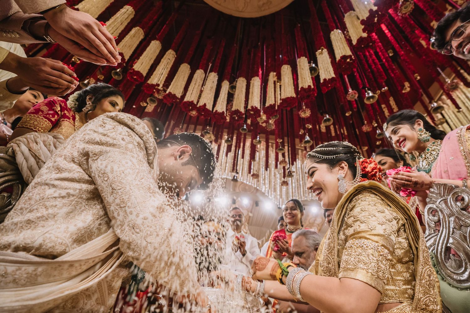 Bride and groom laugh during vibrant Indian wedding ceremony under decorative red and gold mandap with flower petals.