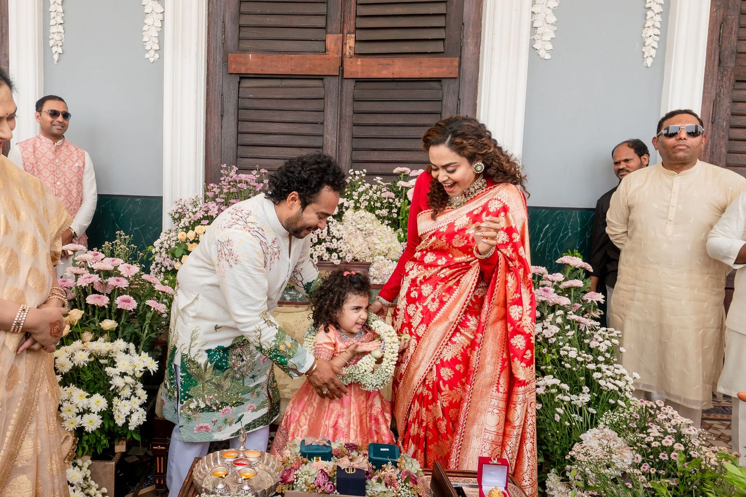 Indian wedding ceremony with groom in white sherwani bowing to flower girl, bride in red saree standing nearby.