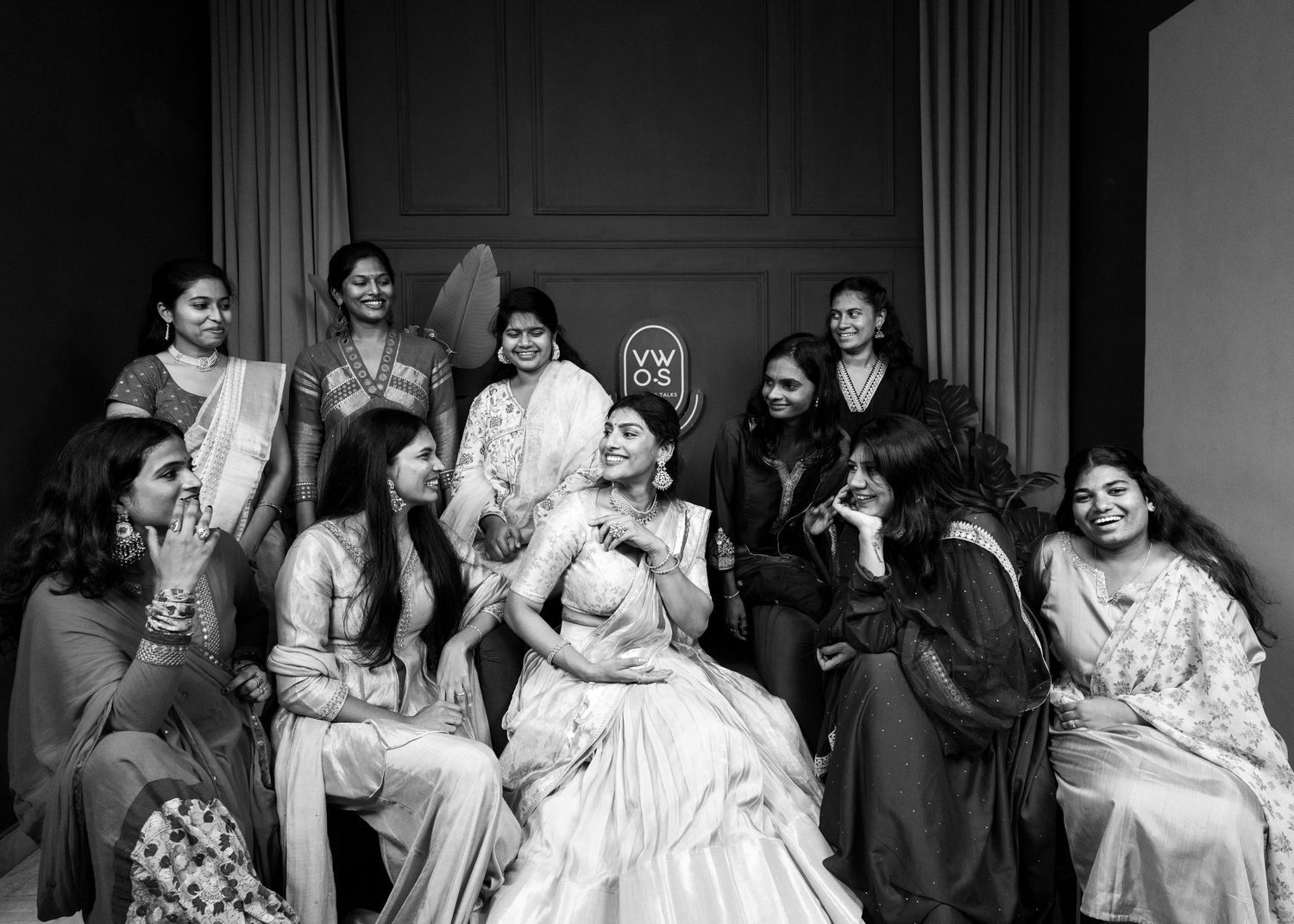 Black and white photo of a bride in lehenga surrounded by laughing bridesmaids at an Indian wedding celebration.