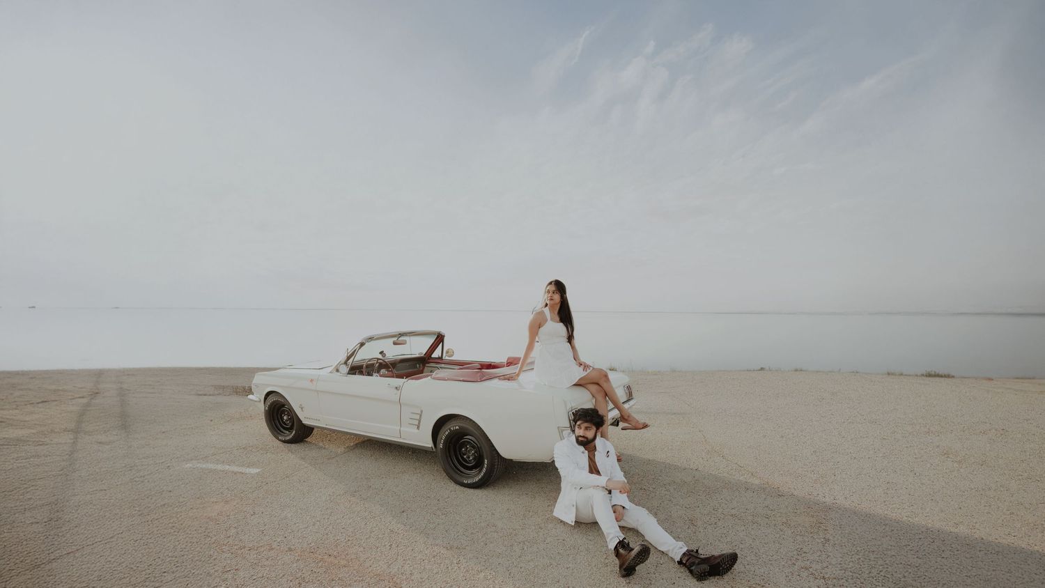 Couple poses with a white vintage Ford Mustang convertible on a sandy beach, woman sitting on hood, man kneeling beside her.