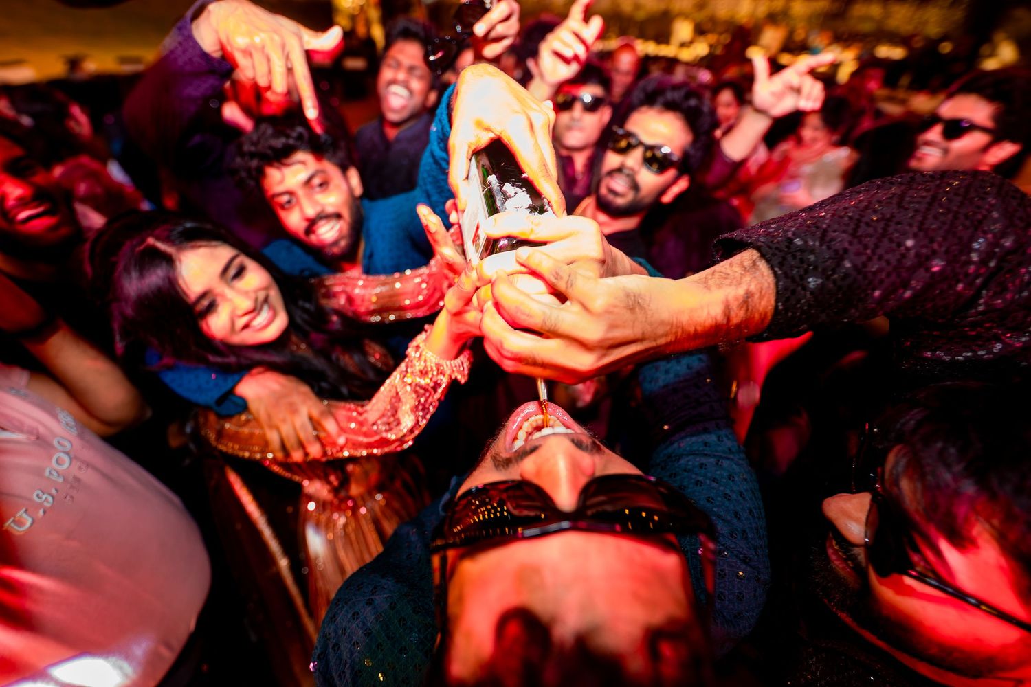 A lively group of friends at a party clinking bottles together in a celebratory toast under colorful lights.