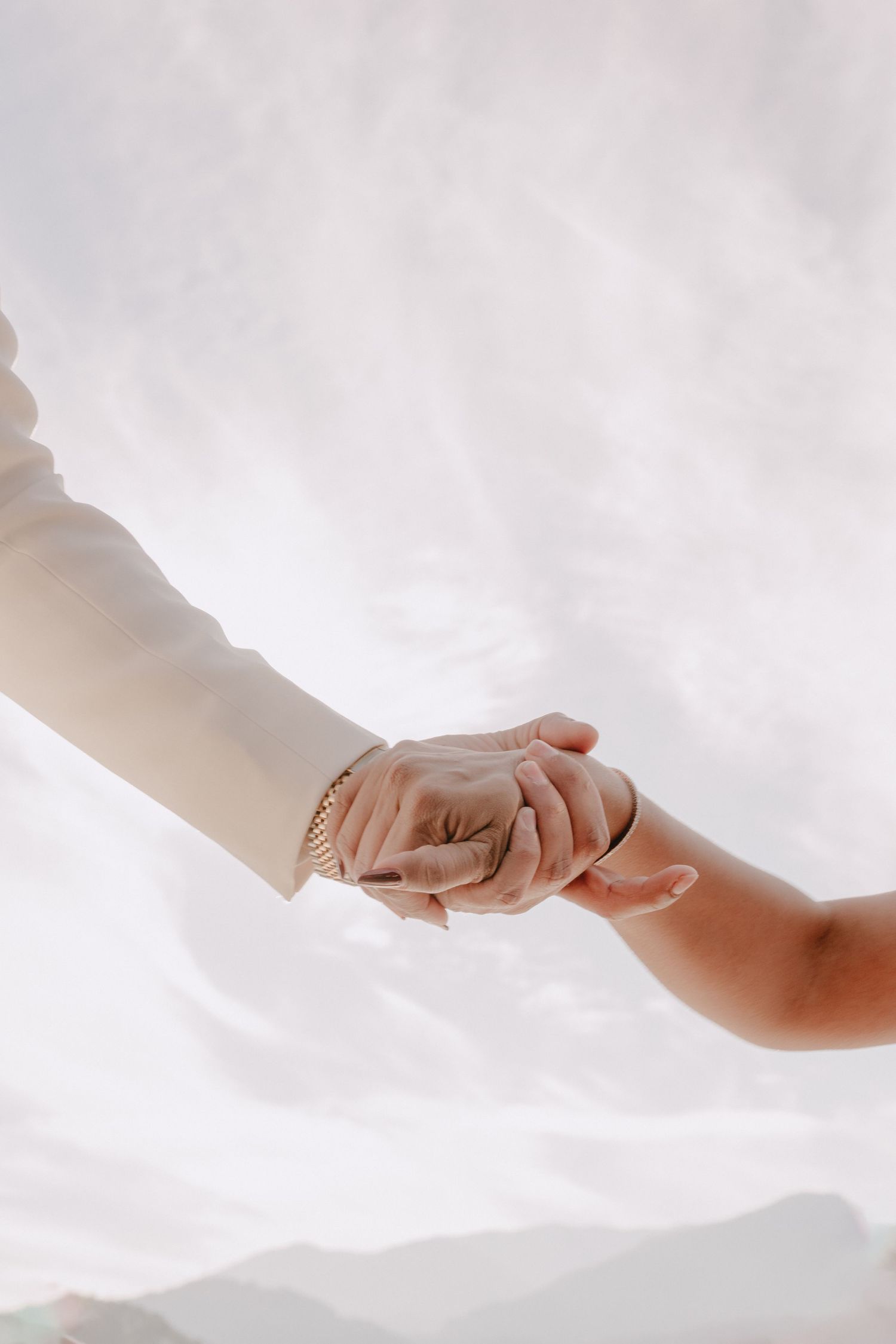 Close-up of bride and groom holding hands, woman wearing gold bracelet against soft white background.