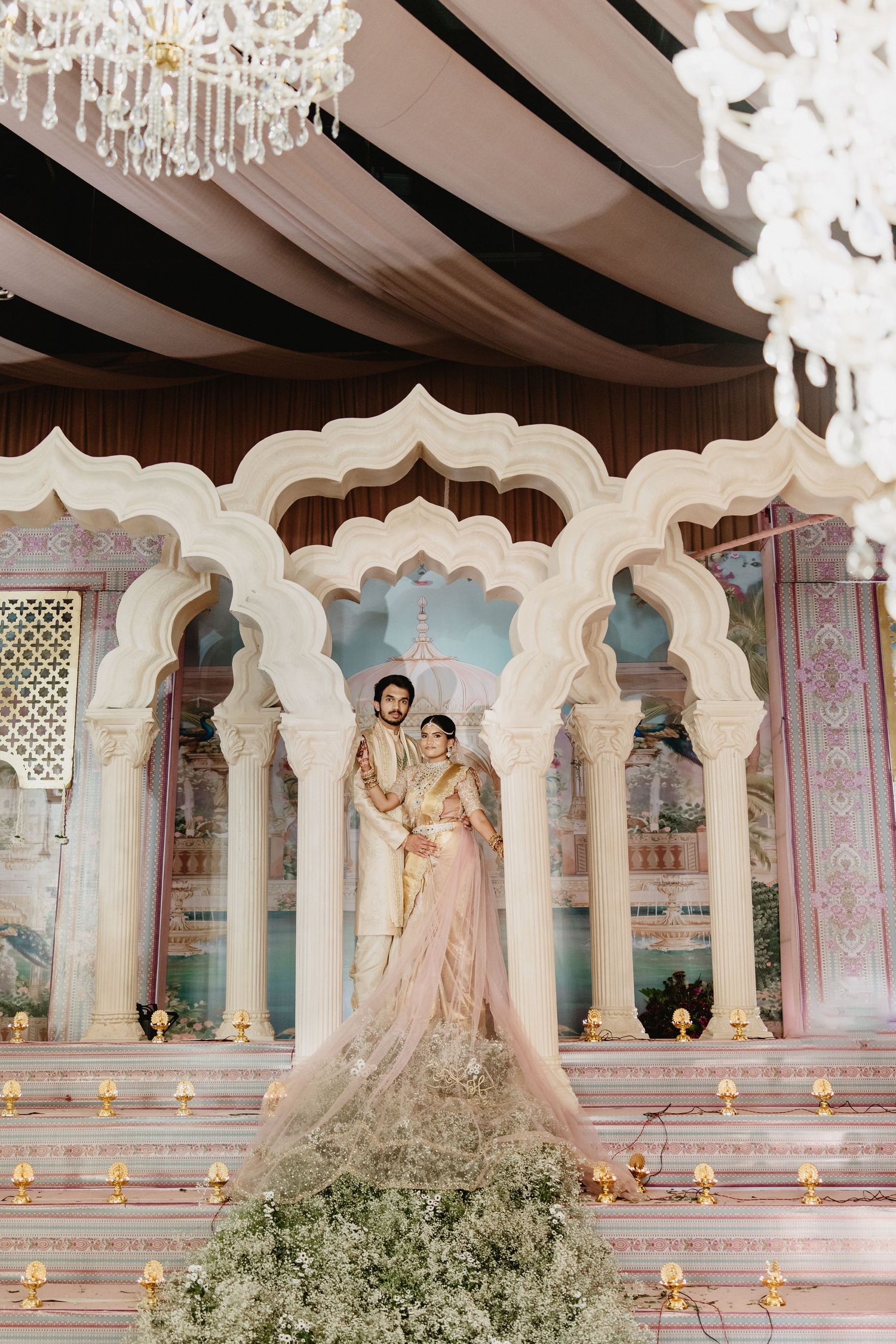 Bride and groom stand together under white Mughal-arch mandap with candles and white florals at elegant Indian wedding.