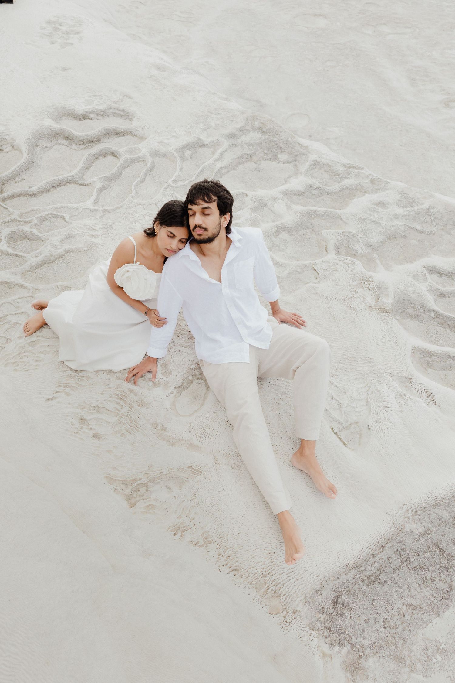 Couple in white outfits reclines together on white sandy beach from aerial view.