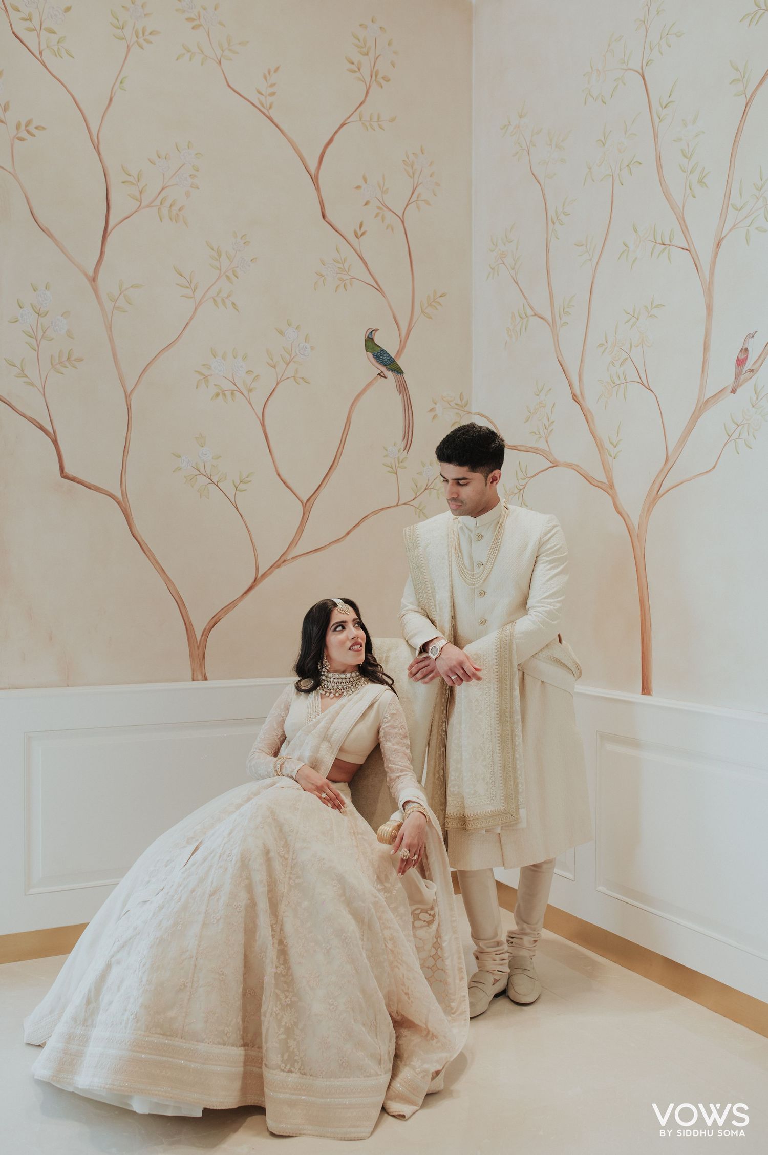 Bride in ivory lehenga sits beside groom in cream sherwani against elegant floral mural backdrop at Indian wedding.