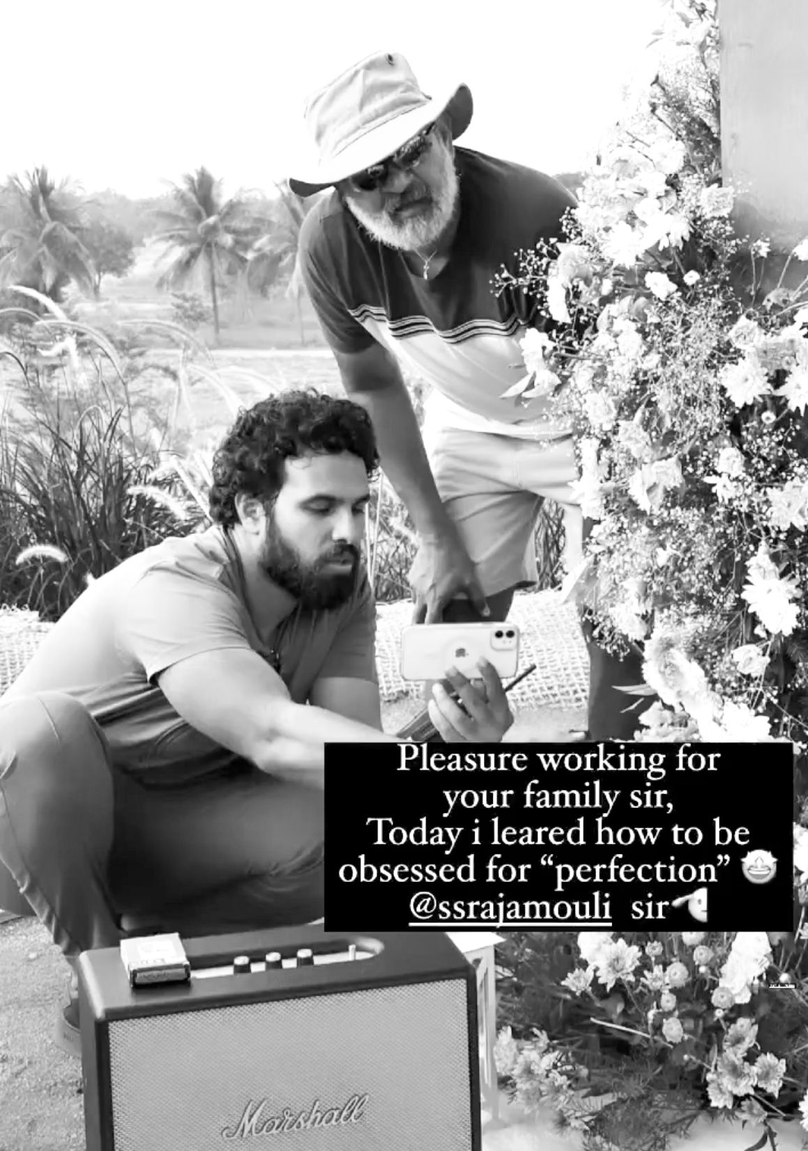 Black and white photo of two men working together near flowers, with a Marshall amp, tagged @ssrajamouli_sir.