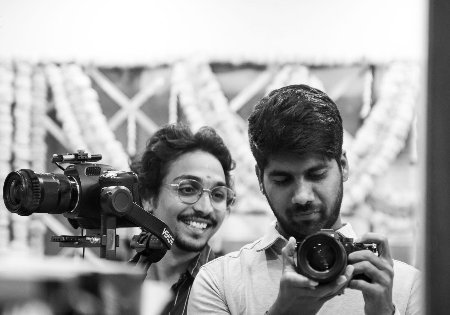 Two young men with cameras smile in a black and white photo at an outdoor event.