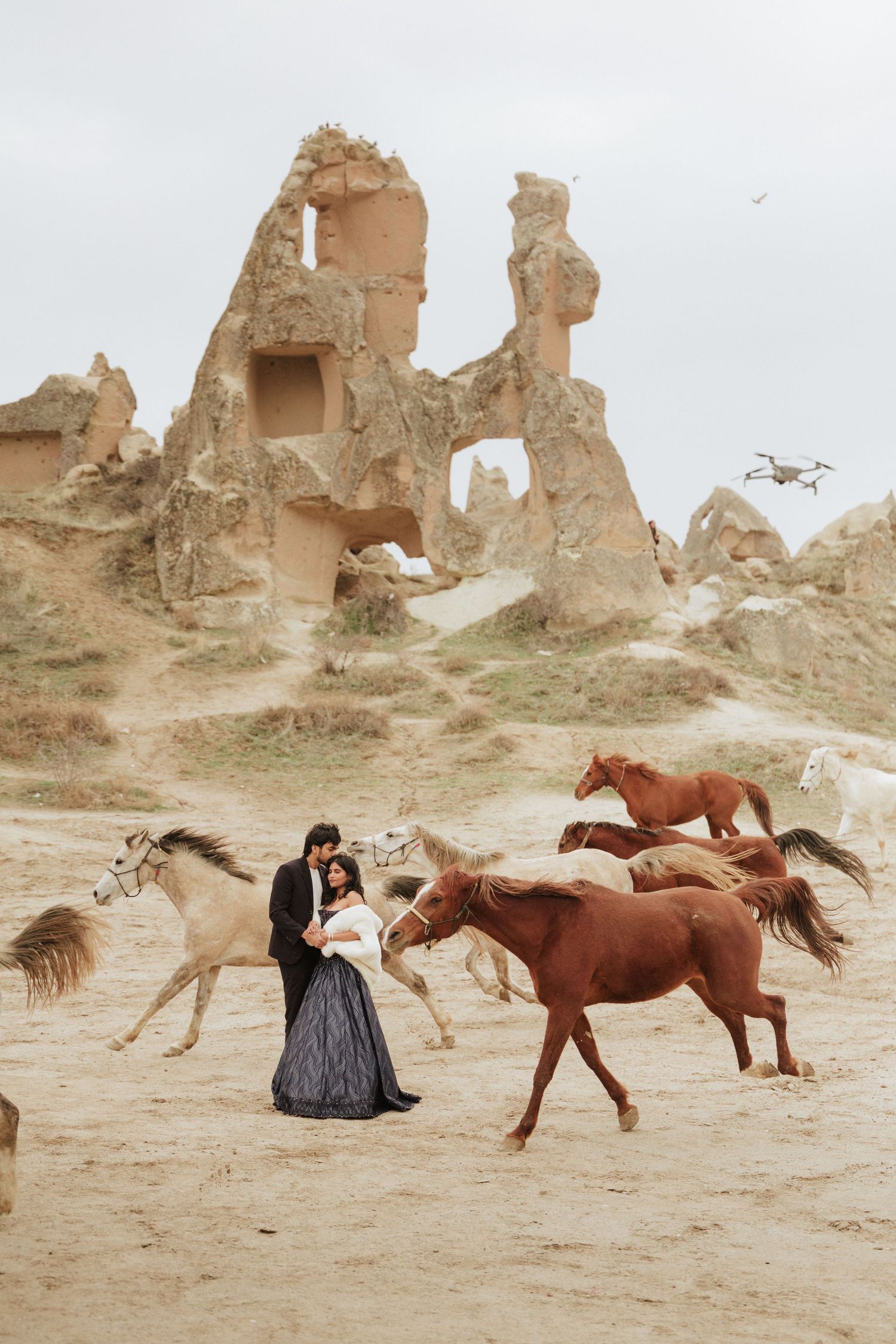 Couple in formal attire embracing as horses gallop around them near Cappadocia rock formations, with a drone flying overhead.