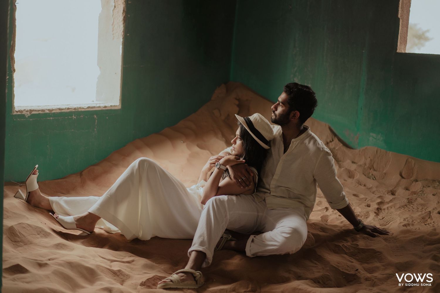 Couple in white outfits sitting on sand dunes inside a room with green walls for a romantic photoshoot.