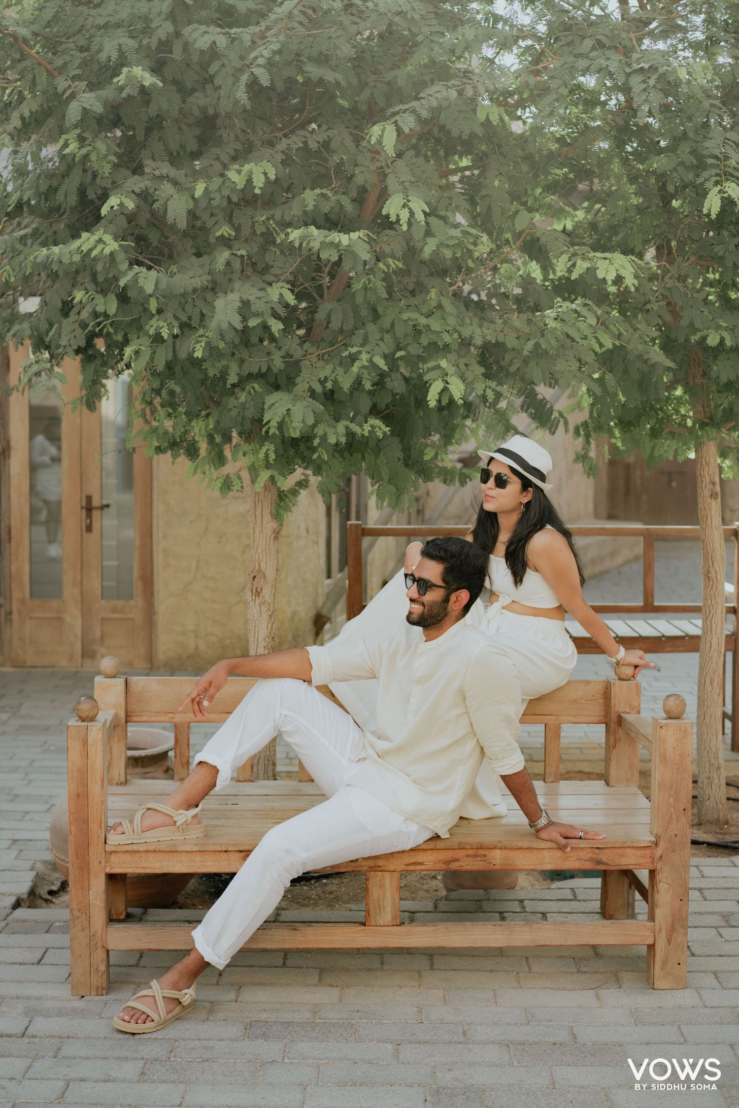 Couple in matching white outfits posing on a wooden bench outdoors, with lush green trees in the background.
