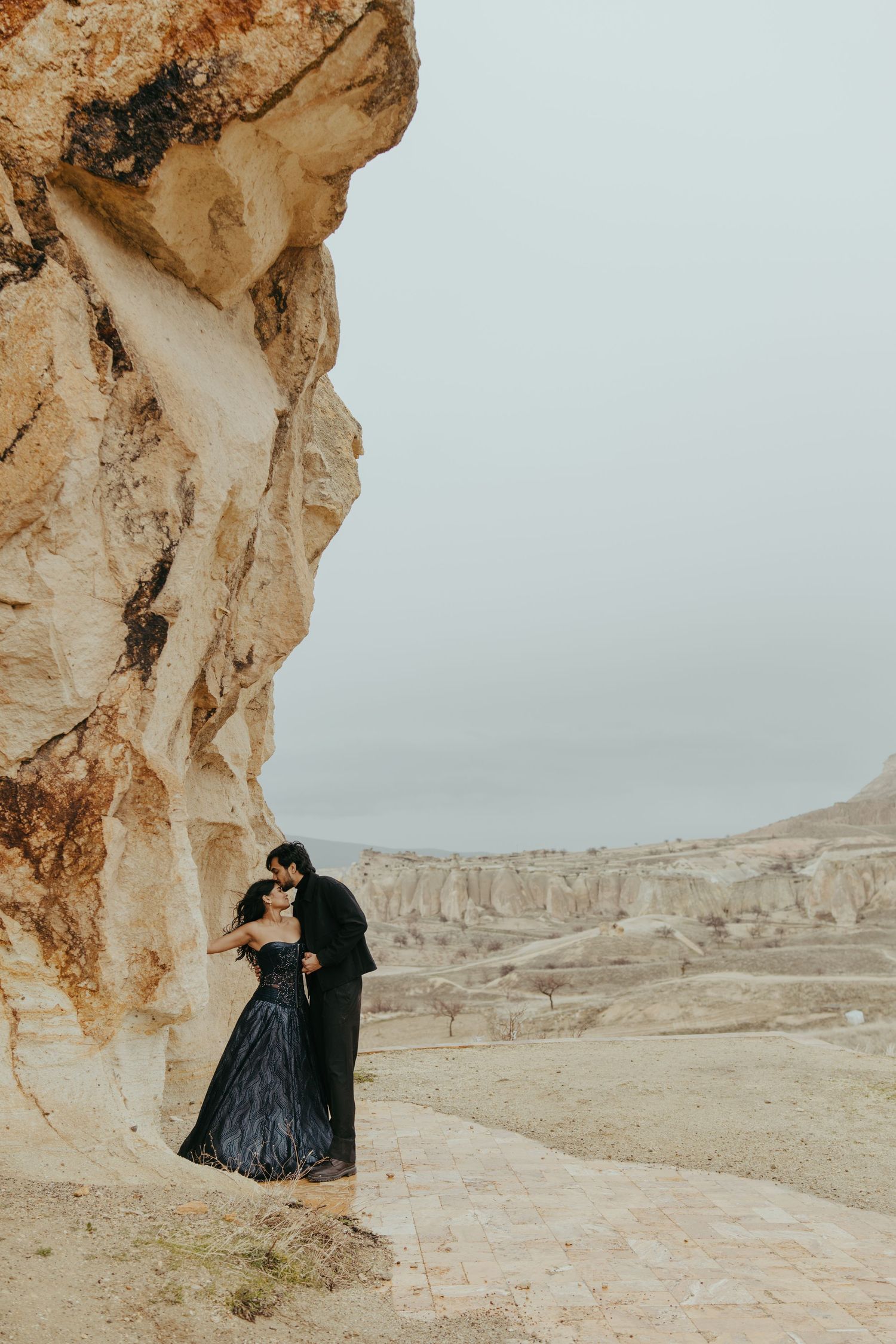 Romantic couple in black wedding attire embracing beside ancient rock formations in Cappadocia, Turkey.